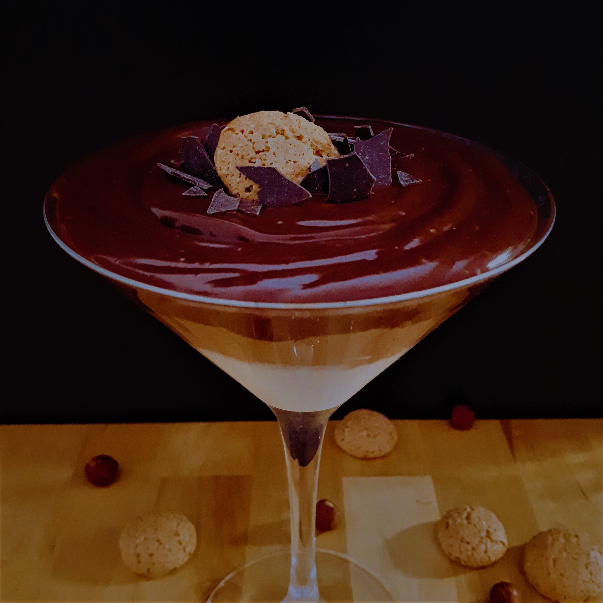 Martini pohár naplnený domácim Monte krémom z troch rôznofarebných vrstiev dozdobený sušienkou Amaretti a kúskami horkej čokolády na drevenom stole okolo rozsypané Amaretti sušienky a lieskové orechy.
