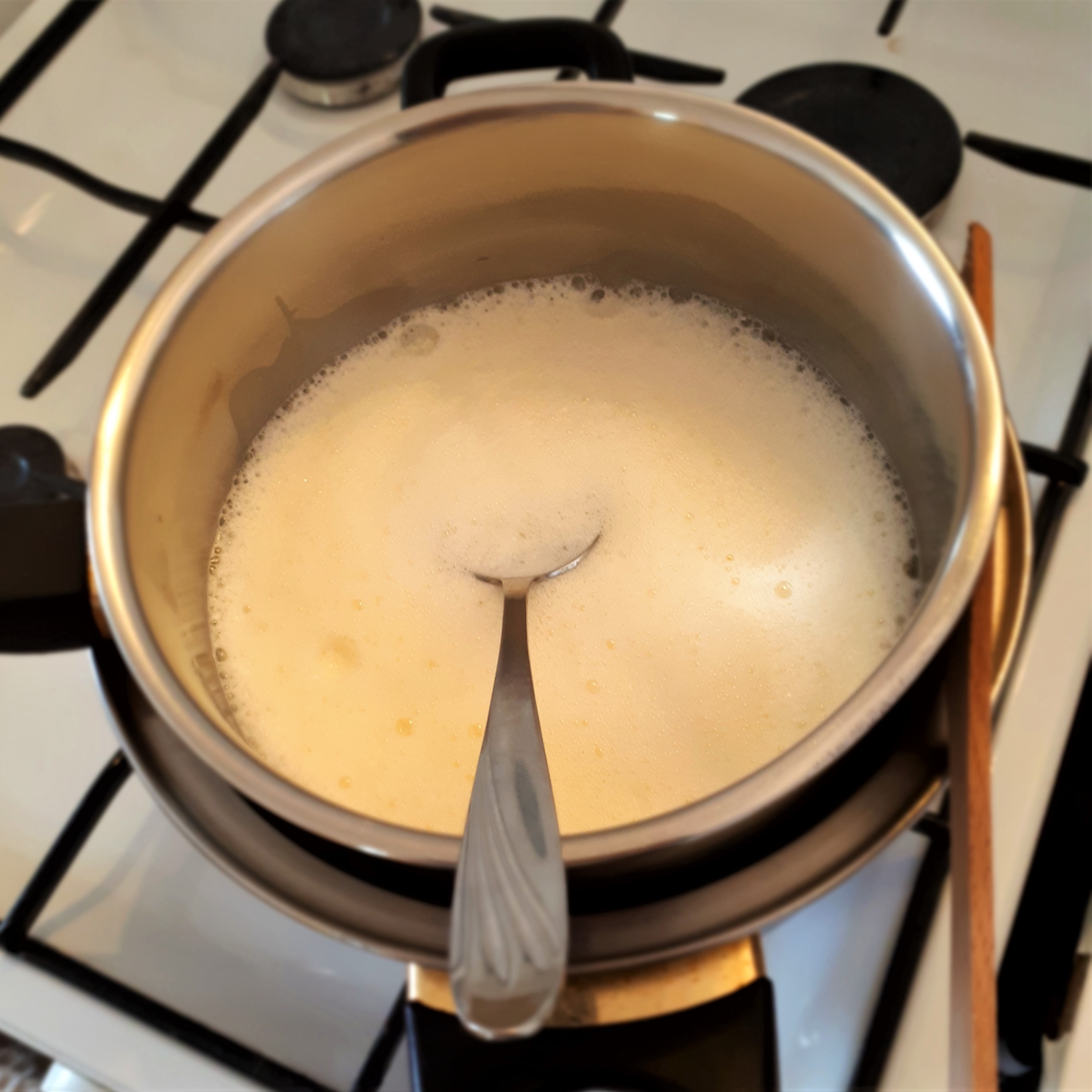 Menší hrniec s krémom na Crème brûlée a polievkovou lyžicou vložený vo väčšom hrnci s vriacou vodou z jednej strany podopretý drevenou varechou na sporáku.