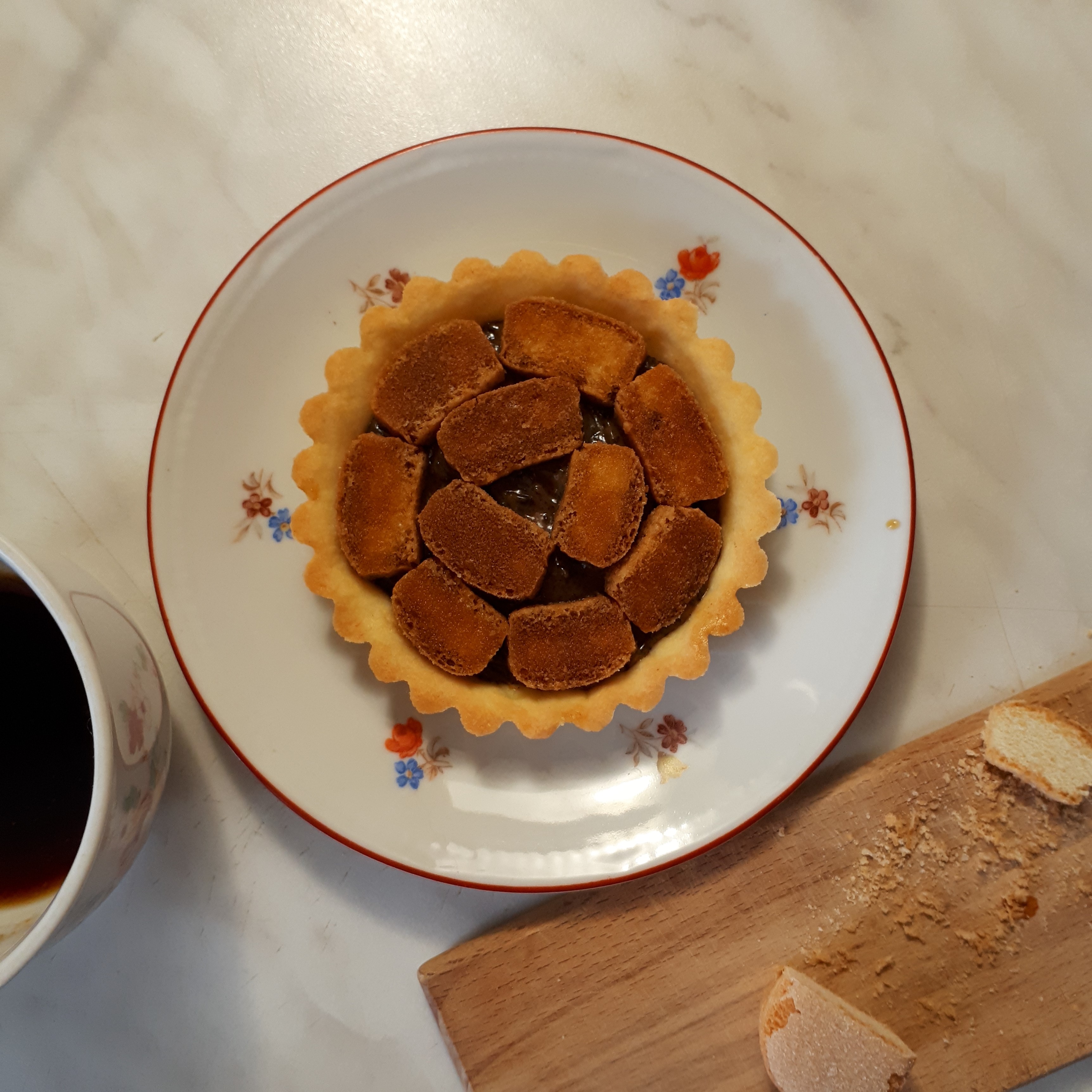 Tartaletka zo svetlého krehkého cesta s kávovou ganache nakrájané cukrárske piškóty namočené v káve biely tanierik s červeným okrajom a drobnými kvietkami hrnček s kávou drevená doska na krájanie pokrájaná cukrárska piškóta