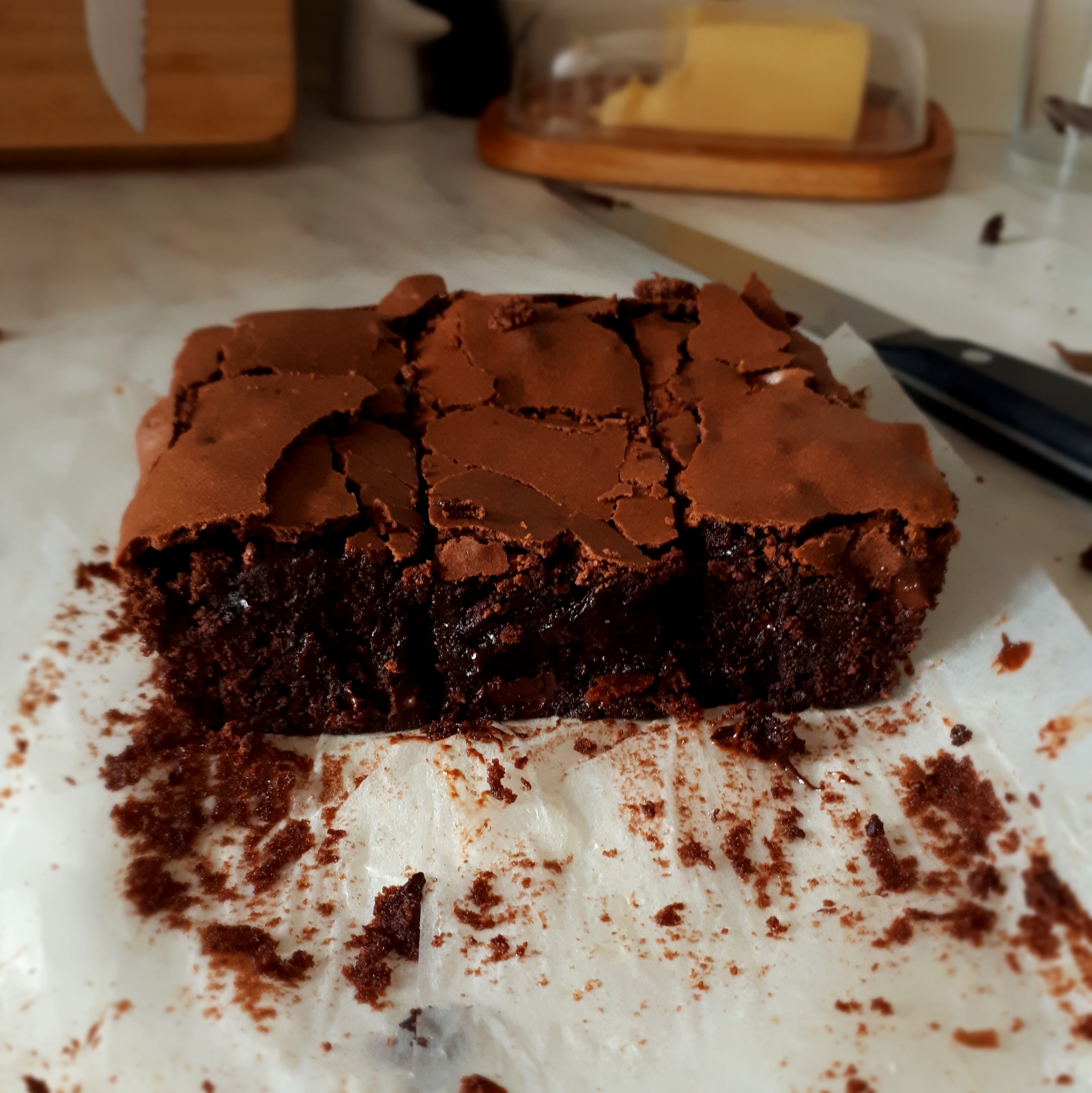 Upečené brownies s chrumkavou kôrkou a roztopenou čokoládou v stede rozložený papier na pečenie nôž maslo v maselničke