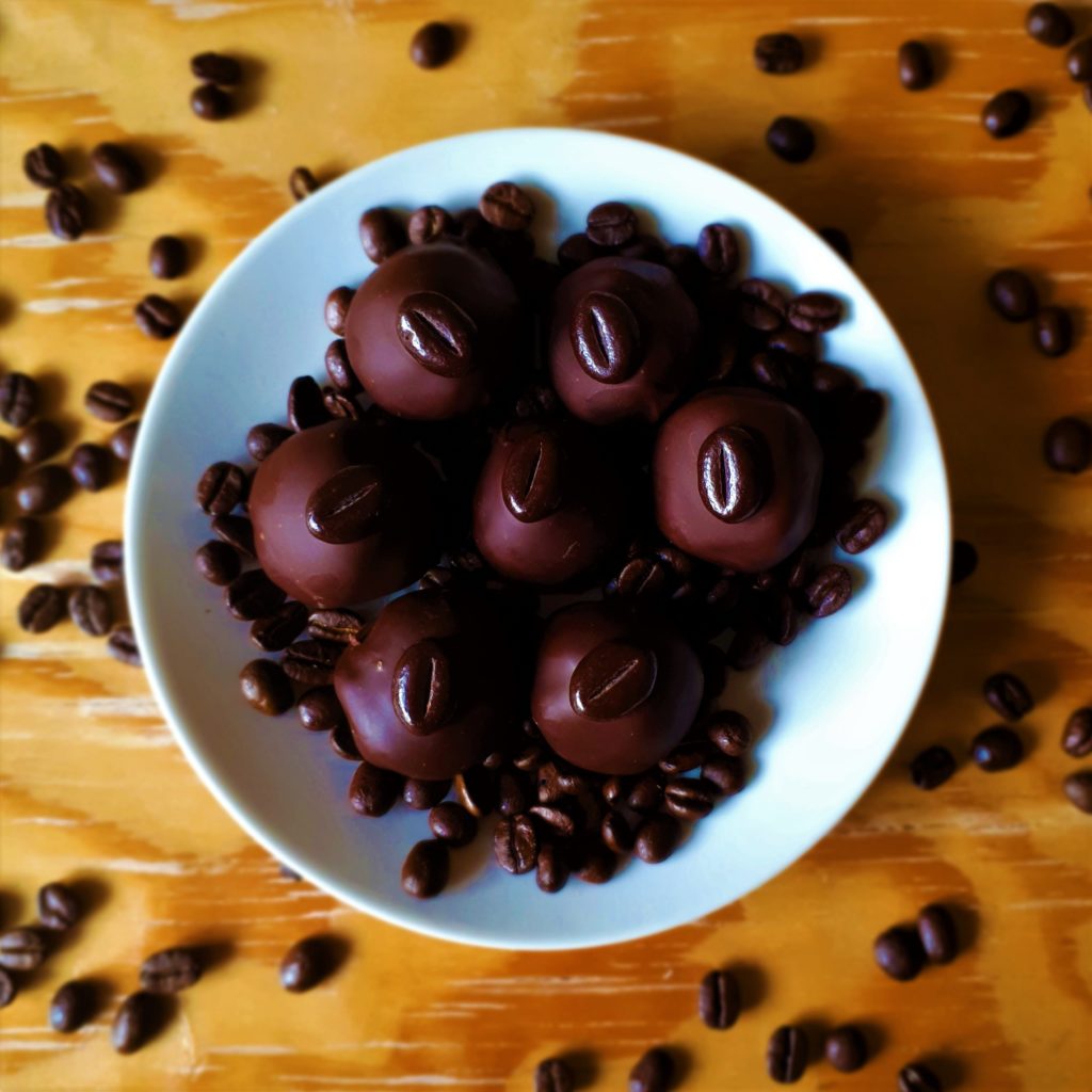 Guľôčky z čokoládovo-kávovej ganache v poleve z tmavej čokolády na bielom tanieri na drevenom stole obsypané kávovými zrnami