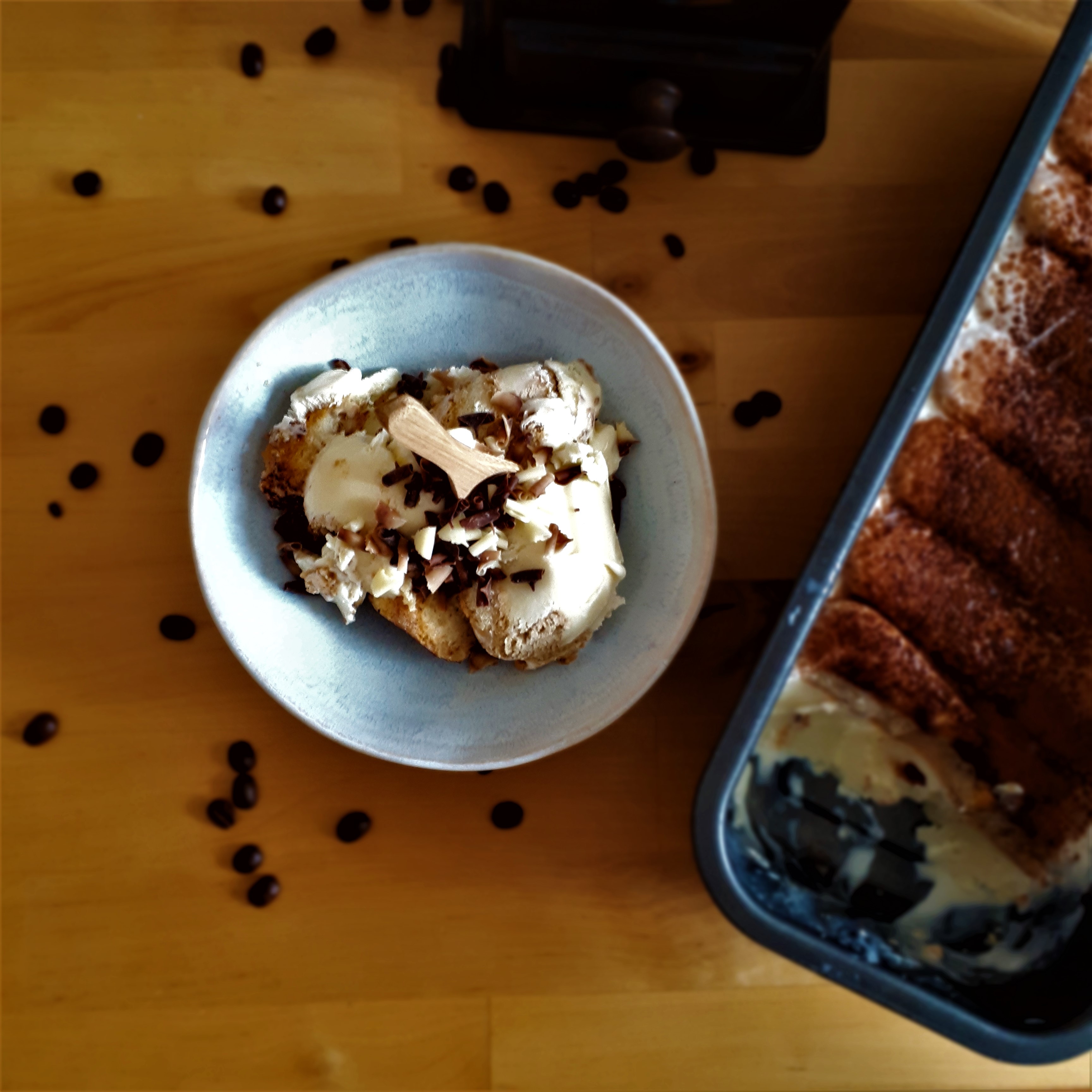 Svetlo-modrá miska s tiramisu zmrzlinou s kúskami cukárskych piškótov posypaná hoblinkami z horkej, mliečnej a bielej čokolády drevená lyžička vysypané zrnká kávy chlebíková forma s tiramisu zmrzlinou mlynček na kávu