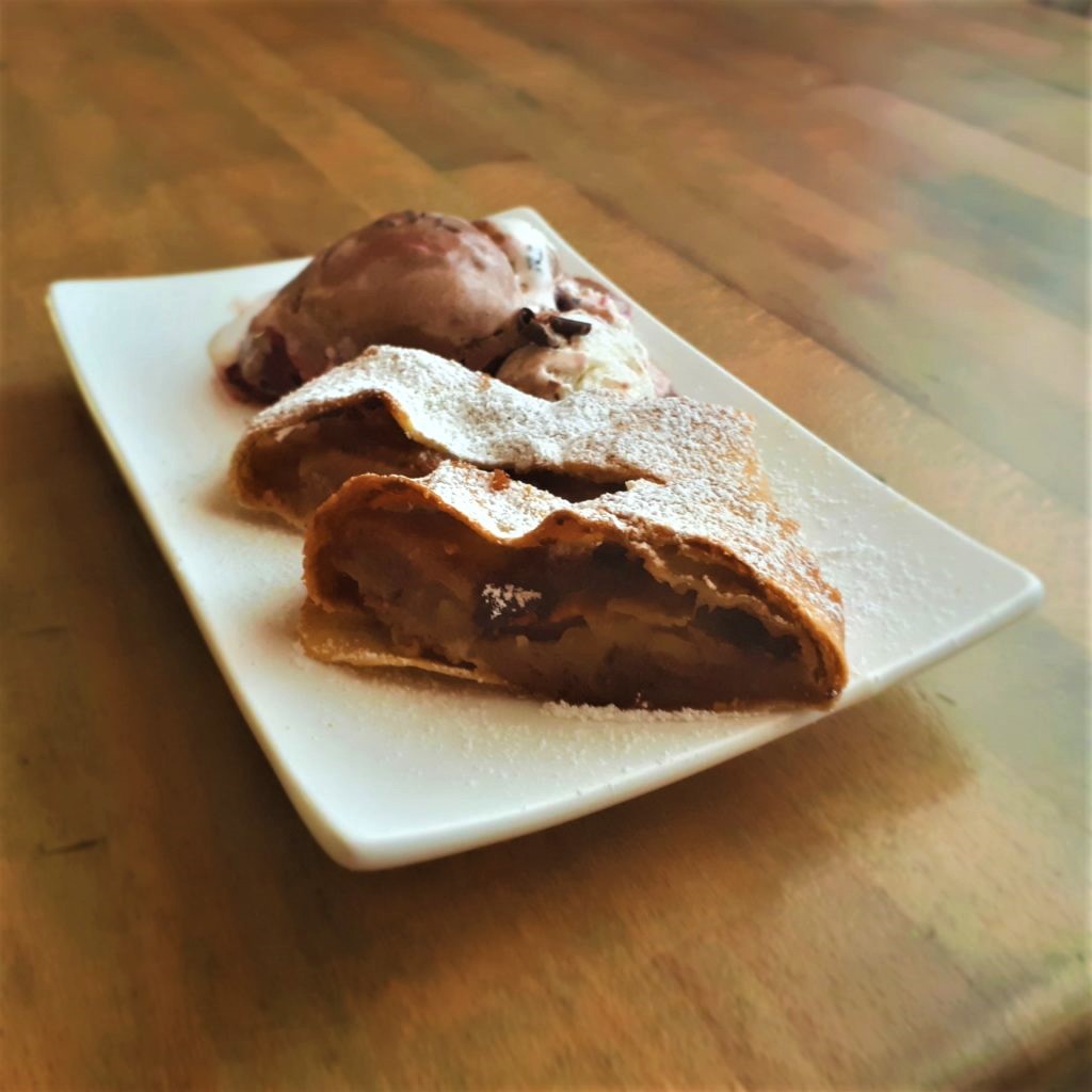 Biely obdĺžnikový tanier s dvoma kúskami jablkovej štrúdle posypané práškovým cukrom čokoládová a vanilková zmrzlina na drevenom stole