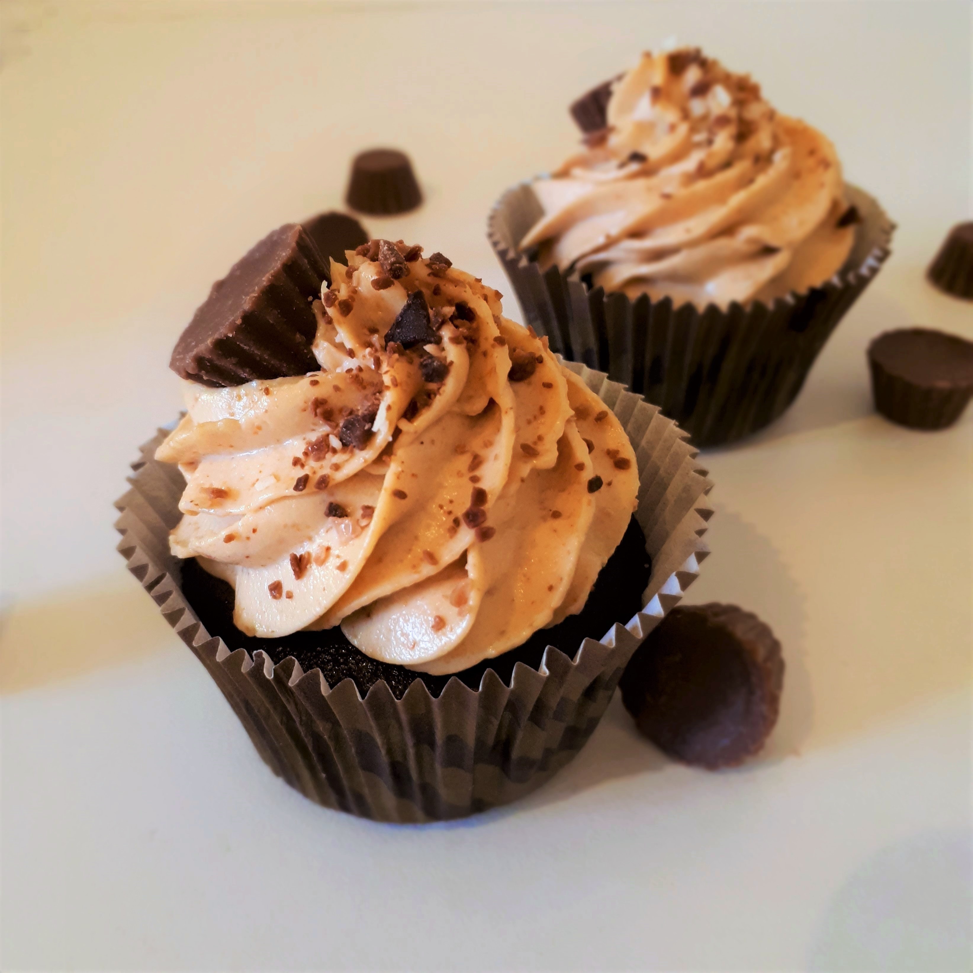 Dva čokoládové cupcakes v hnedom papierovom košíčku krém z arašidového masla posypané nastrúhanou čokoládou s čokoládovým košíčkom na ozdobu okolo rozypané mini čokoládové košíčky na bielom povrcuh