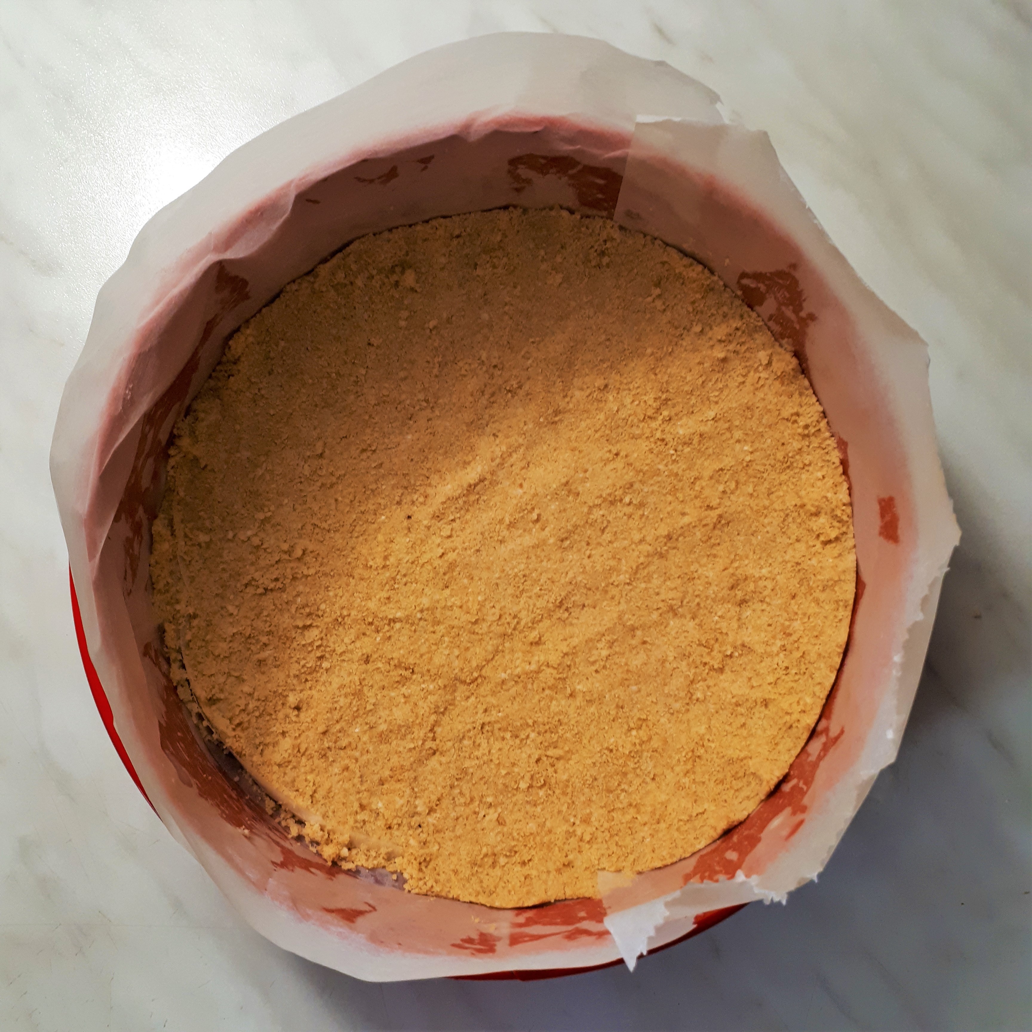 Červená tortová forma vystlaná papierom na pečenie na dne utlačená vrstva sušienok s maslom na cheesecake korpu na mramorovom povrchu