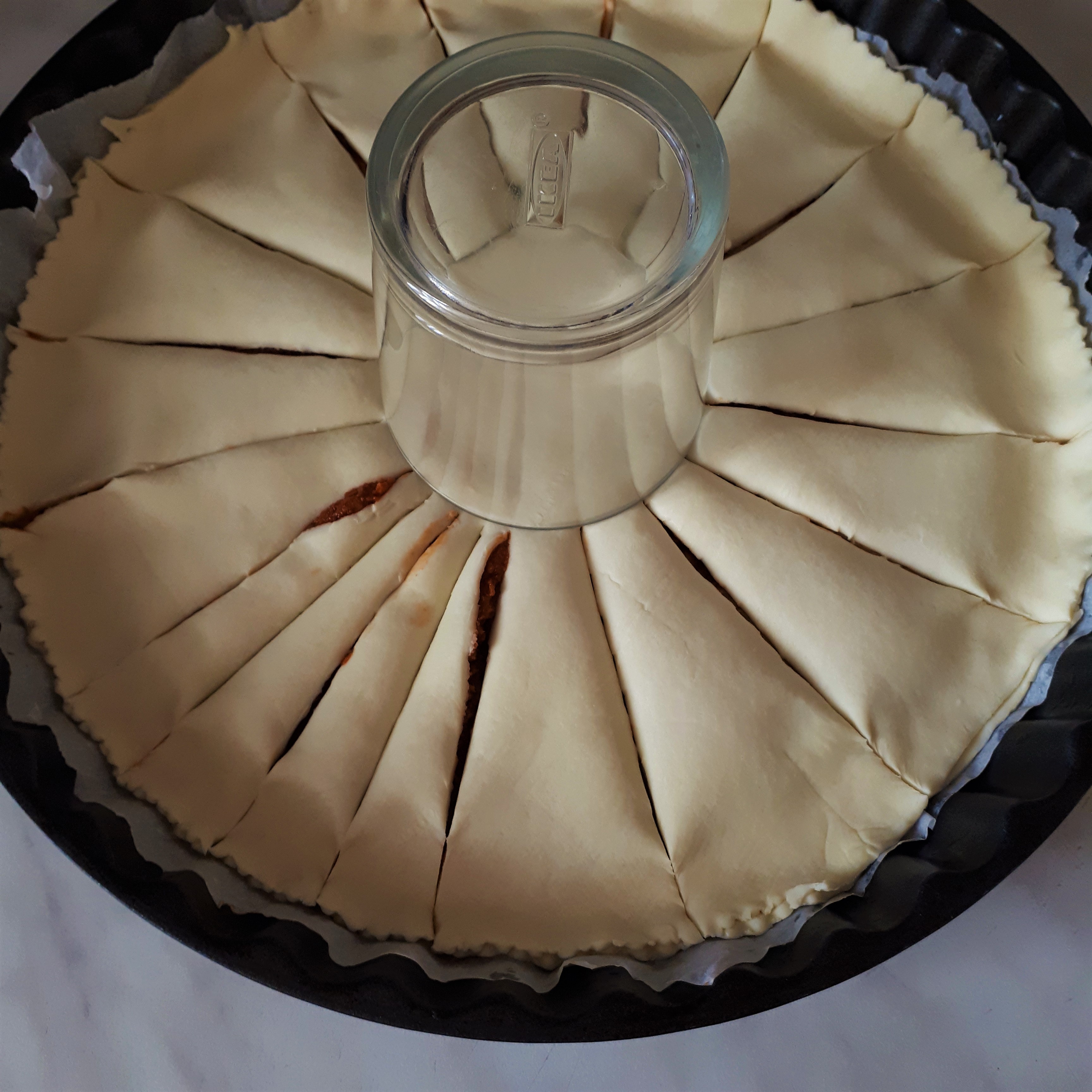 Kruhová forma na koláč vystlaná papierom na pečenie lístkové cesto plnené rajčinovo-olivovou náplňou v strede sklený pohár hore dnom nakrájané pásy cesta okolo pohára na mramorovom povrchu
