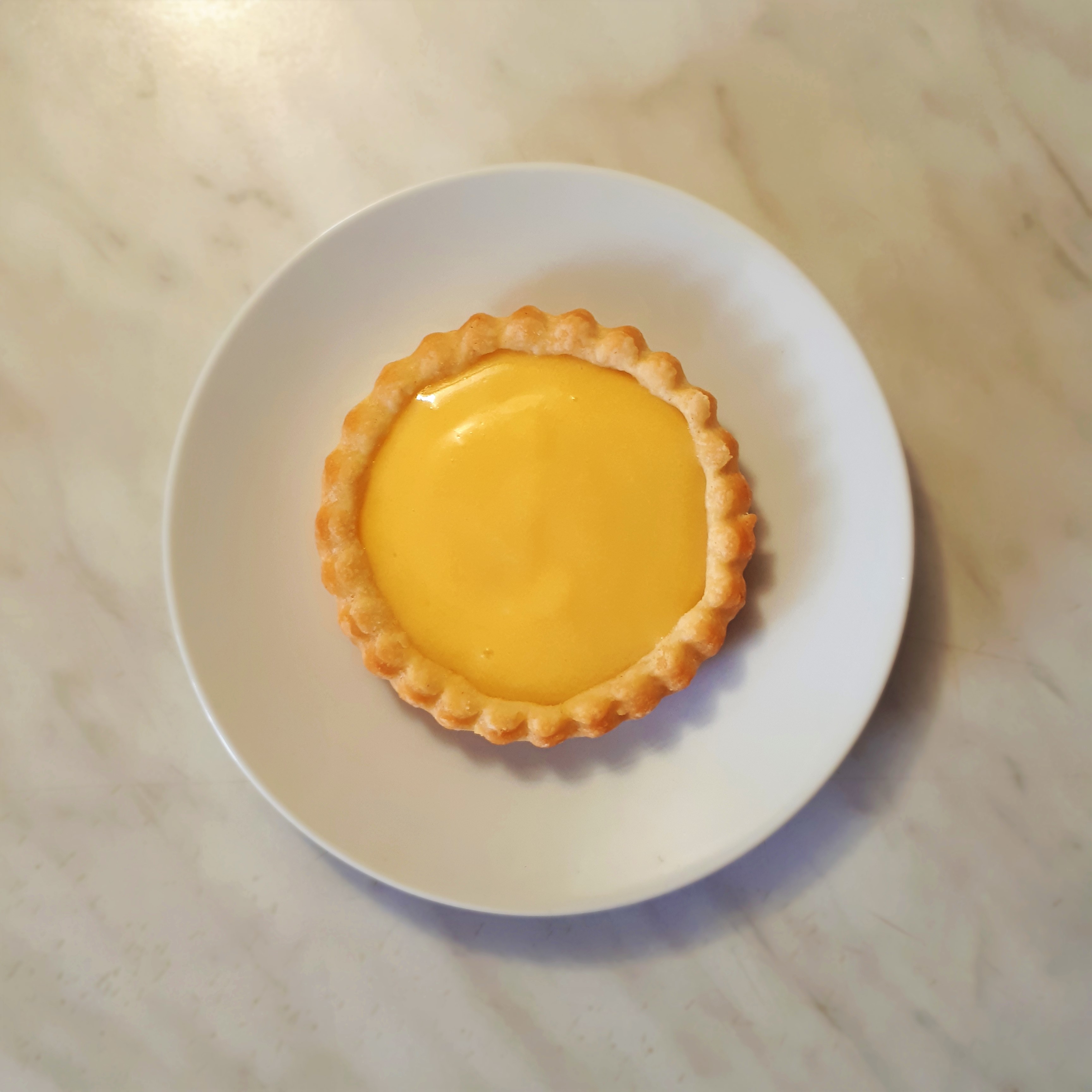 Tartaletka zo svetlého krehkého cesta naplnená citrónovým krémom lemon curd na bieom tanieri mramoroví kuchynskí linka