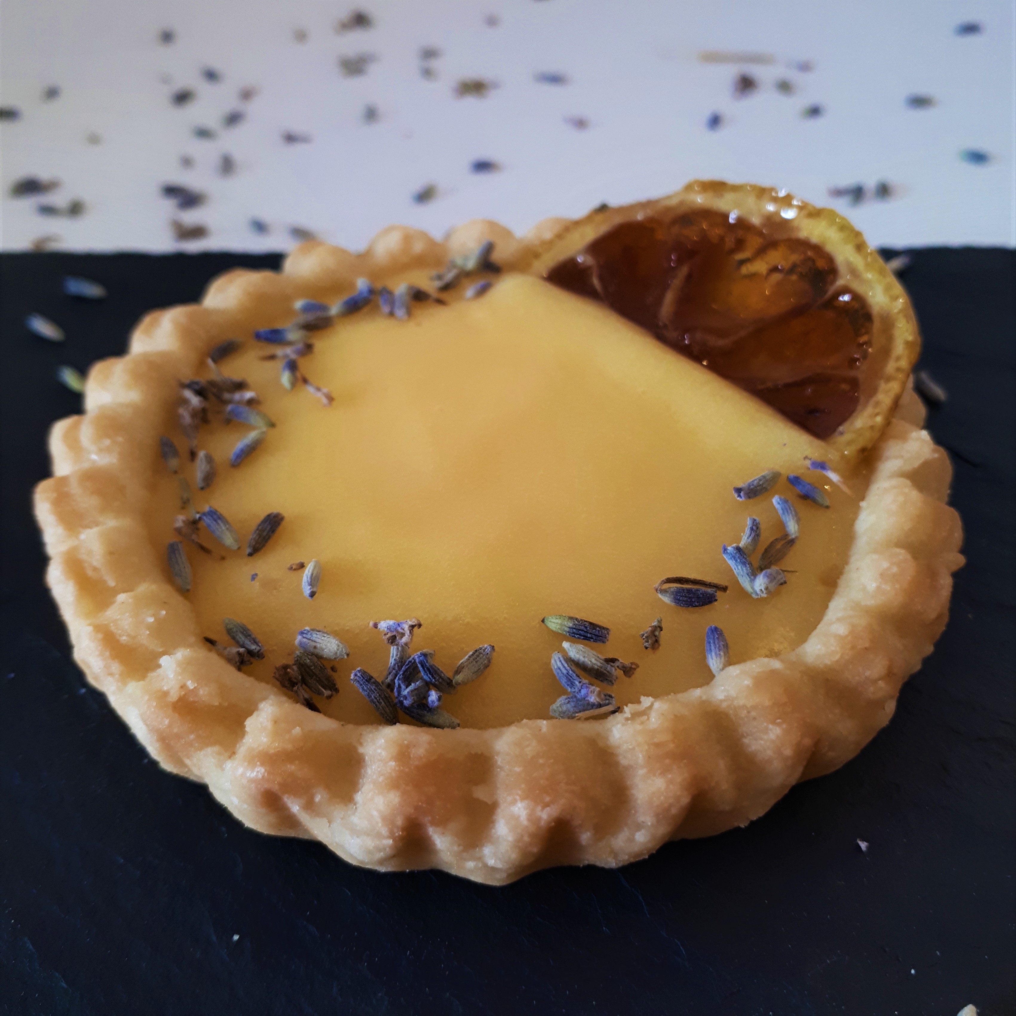 Tartaletka zo svetlého krehkého cesta naplnená citrónovým krémom lemon curd dozdobená plátkom karamelizovaného citróna a sušenou levanduľou na čiernej mramorovej doske na bielom povrchu