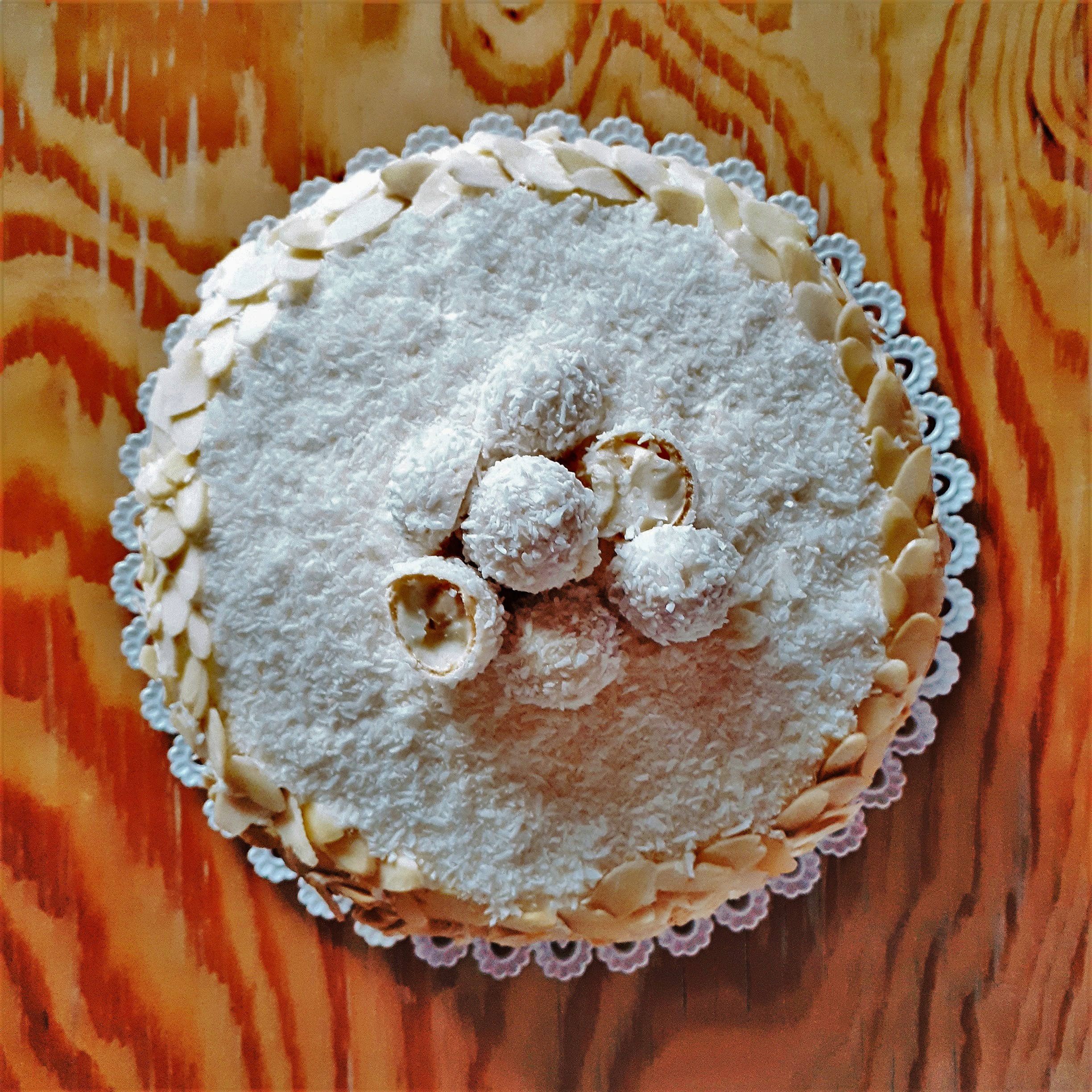 Raffaello torta s kokosom a plátkami mandlí navrchu Raffaello guľôčky na bielej tortovej podložke drevenom stole