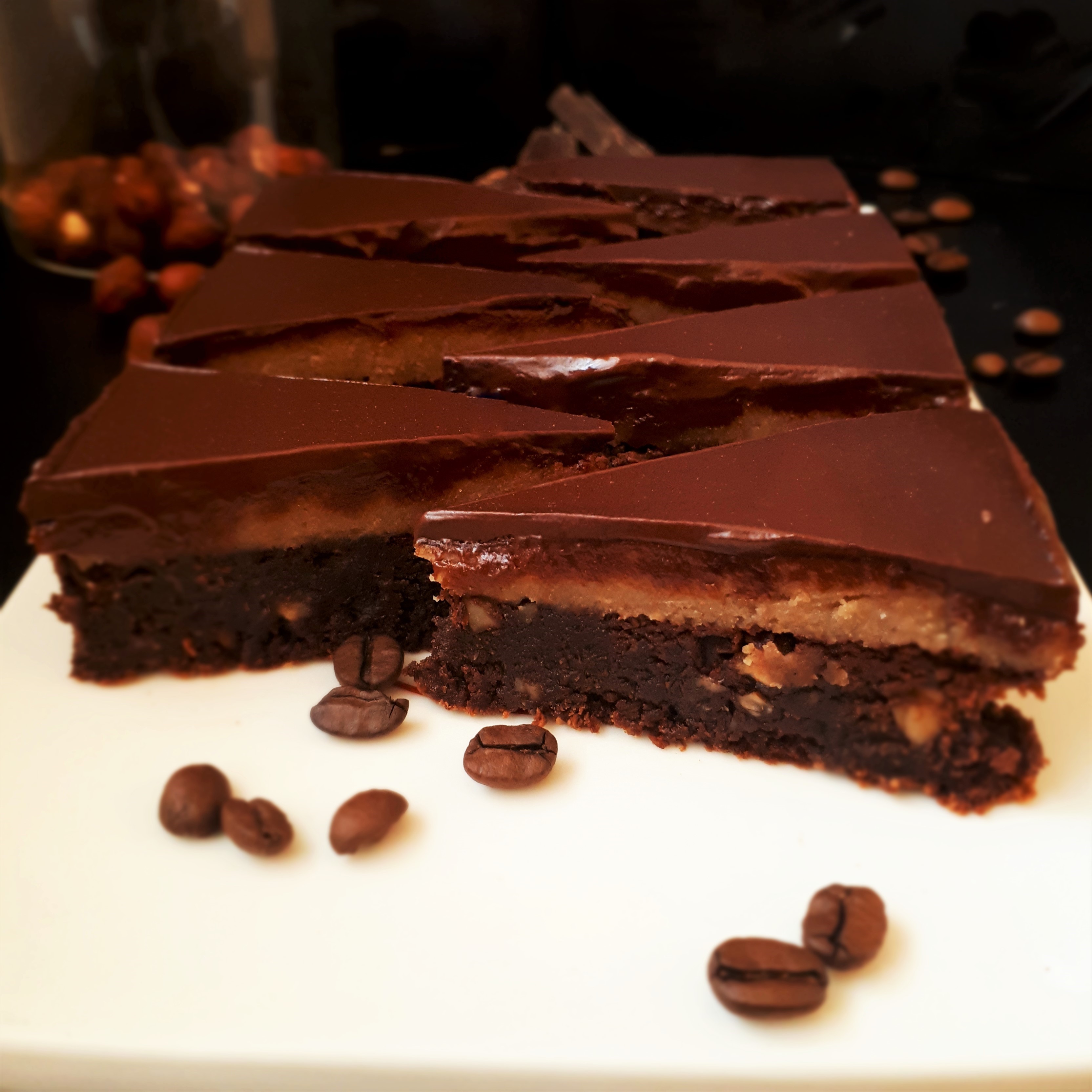 Sedem trojuholníkov brownies poliateho čokoládou na bielom obdĺžnikovom tanieri obsypané kávovými zrnami sklená dóza s lieskovými orechmi kúsky horkej čokolády