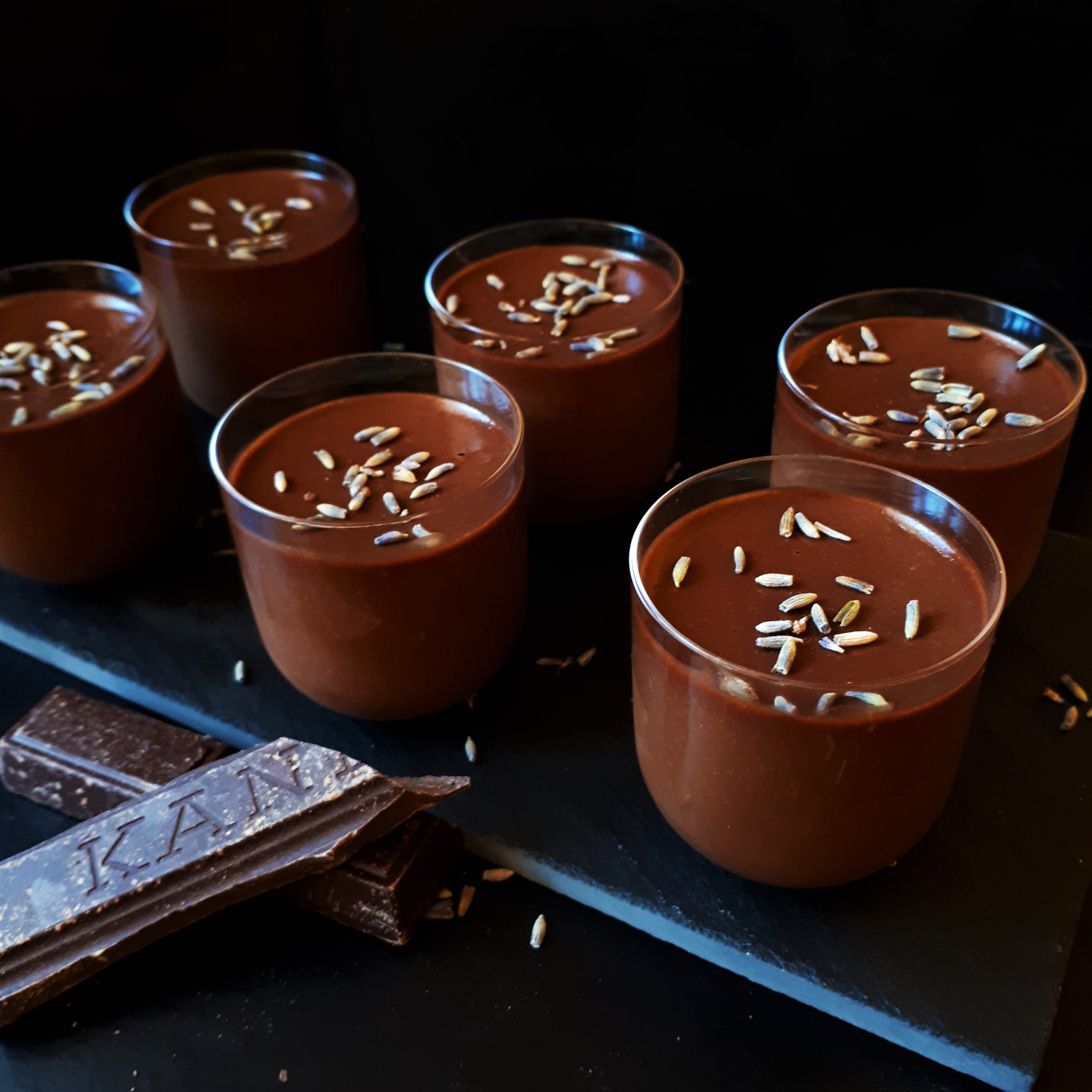 Šesť pohárikov s čokoládovo-levanduľovým krémom posypaných sušenými levanduľovými kvetmi na čiernej kamennej doske kúsok horkej čokolády čiernej pozadie