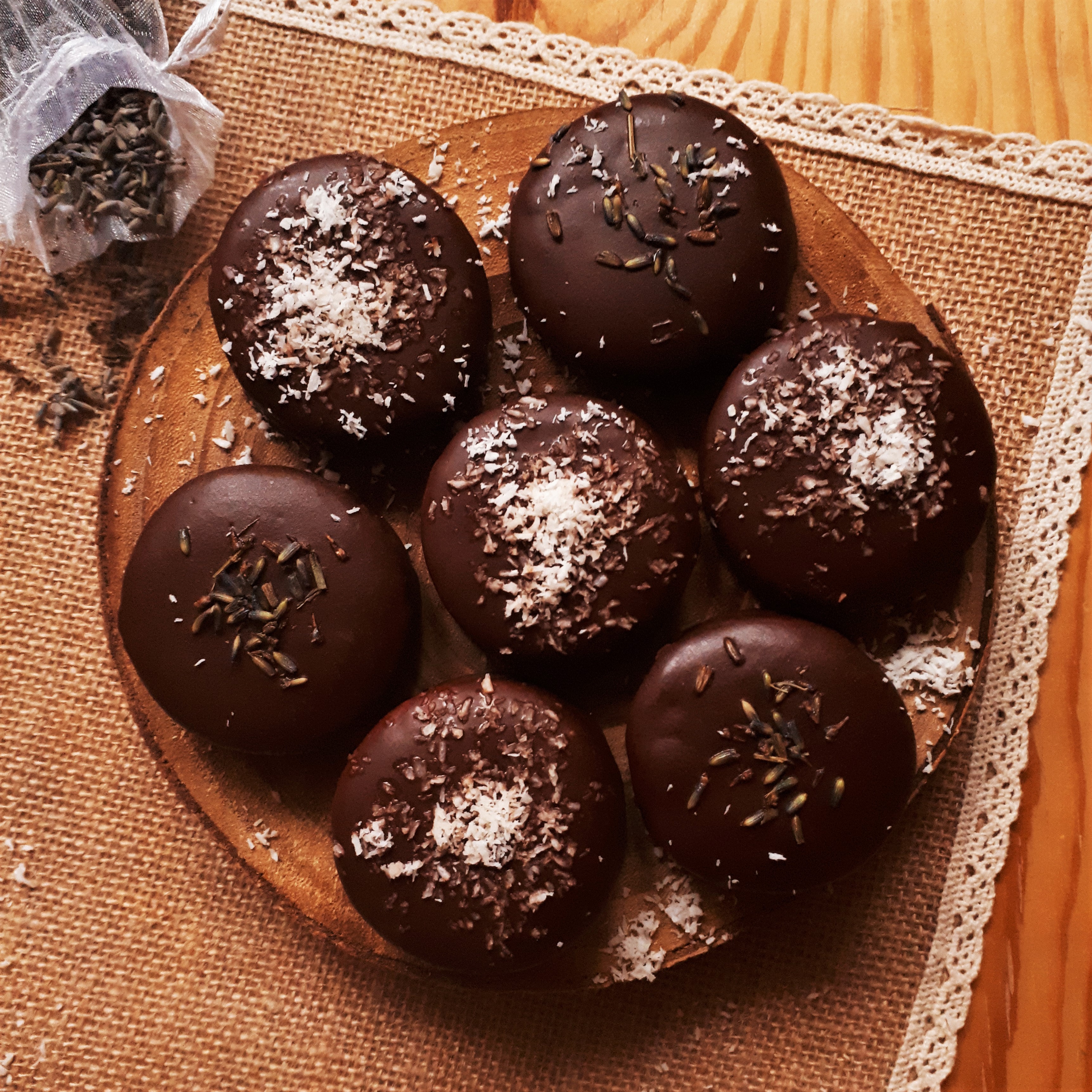 Sedem perníčkov plnených džemom obalených v čokoláde a posypaných kokosom a sušenou levanduľou na drevenom podnose plstenná podložka sáčik s levanduľou a čierno biela kuchynská utierka
