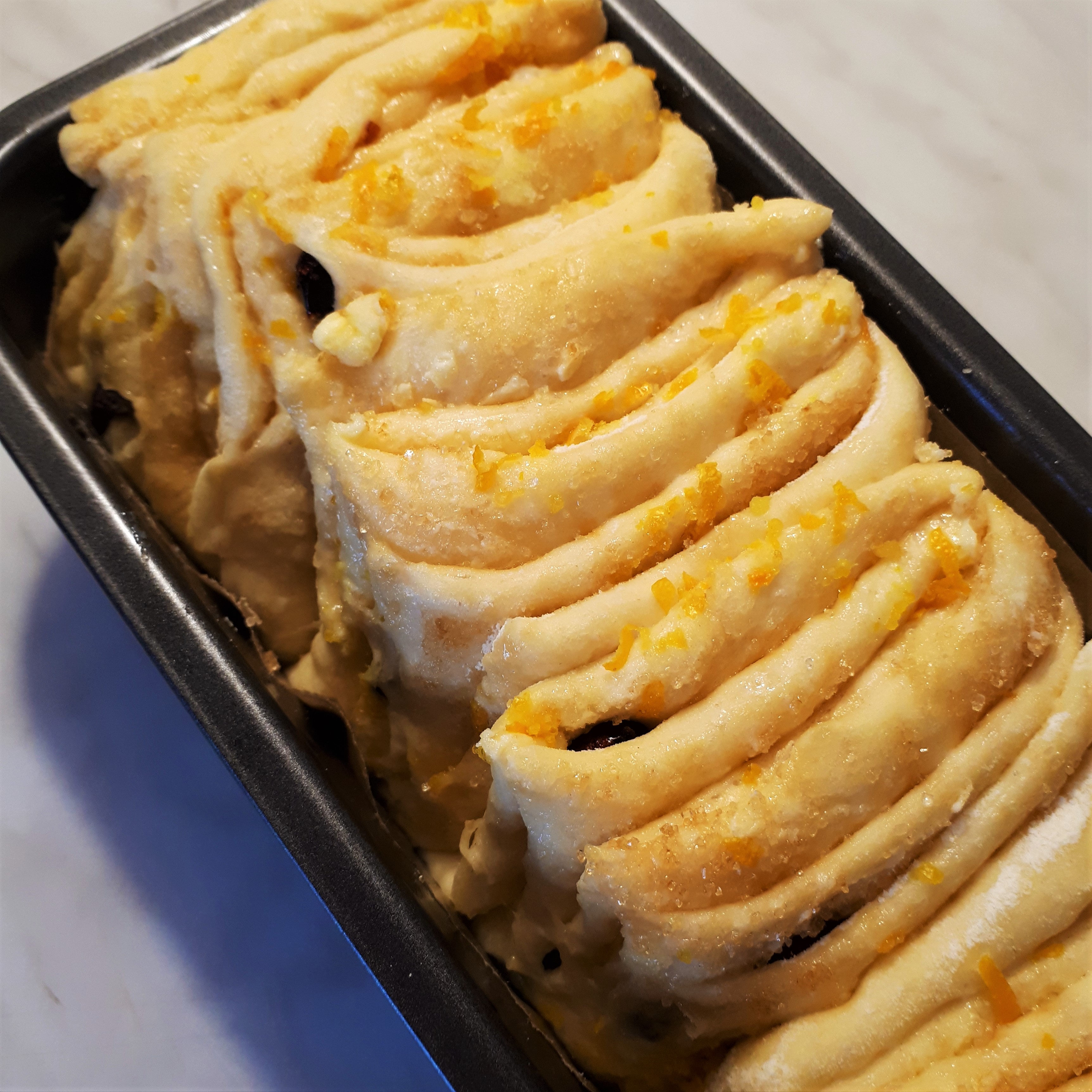 Tmavá forma na chlebík s papierom na pečenie a kysnutým cestom plneným maslom, trstnovým cukrom, strúhanou pomarančovou kôrou a sušenými brusnicami