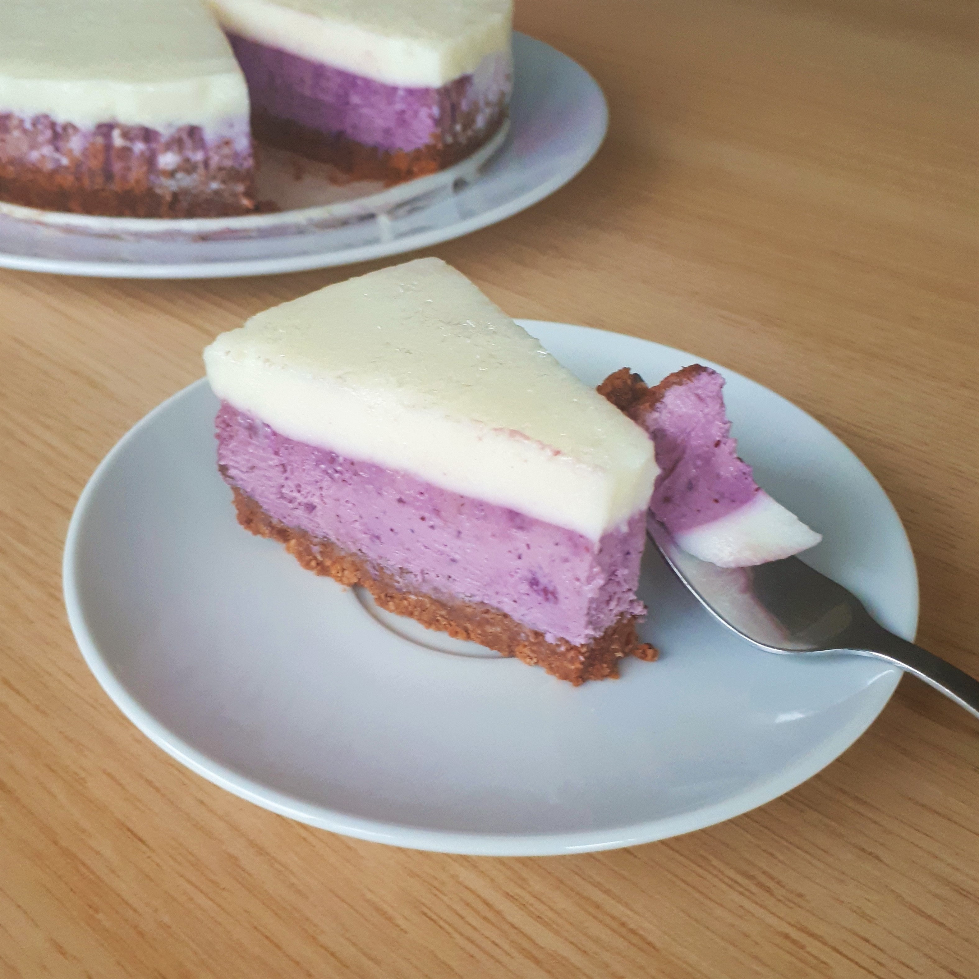 Kúsok dánskeho čučoriedkového cheesecake s bielou smotanovou vrstvou na vrchu posypaný jedlými flitrami na bielom tanieri vidlička s kúskom cheesecake v pozadí zvyšok cheesecake na bielom tanieri na drevenom stole