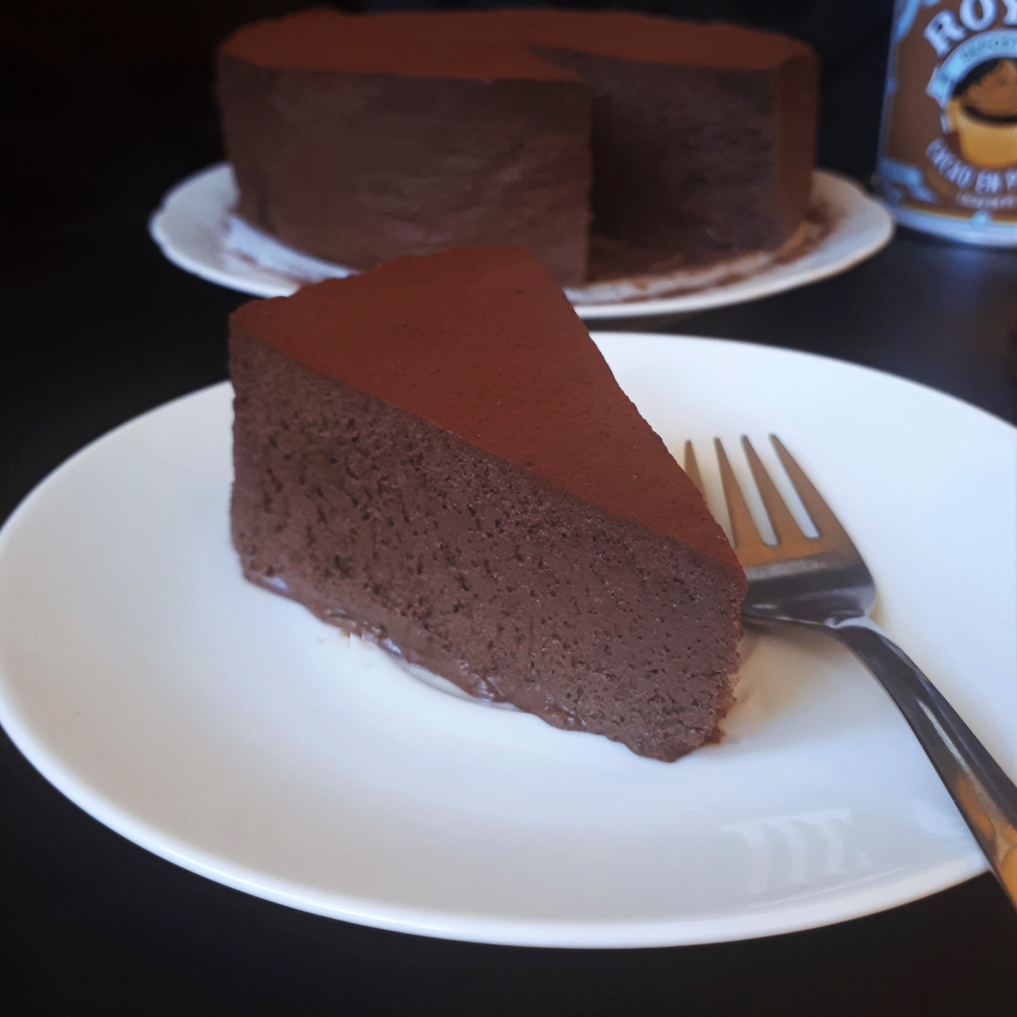 Kúsok penovej čokoládovej torty na bielom tanieri posypanej kakaom antikorová dezertná vidlička na čiernom povrchu v pozadí zvyšok torty na bielom tabnieri a dóza s kakaom