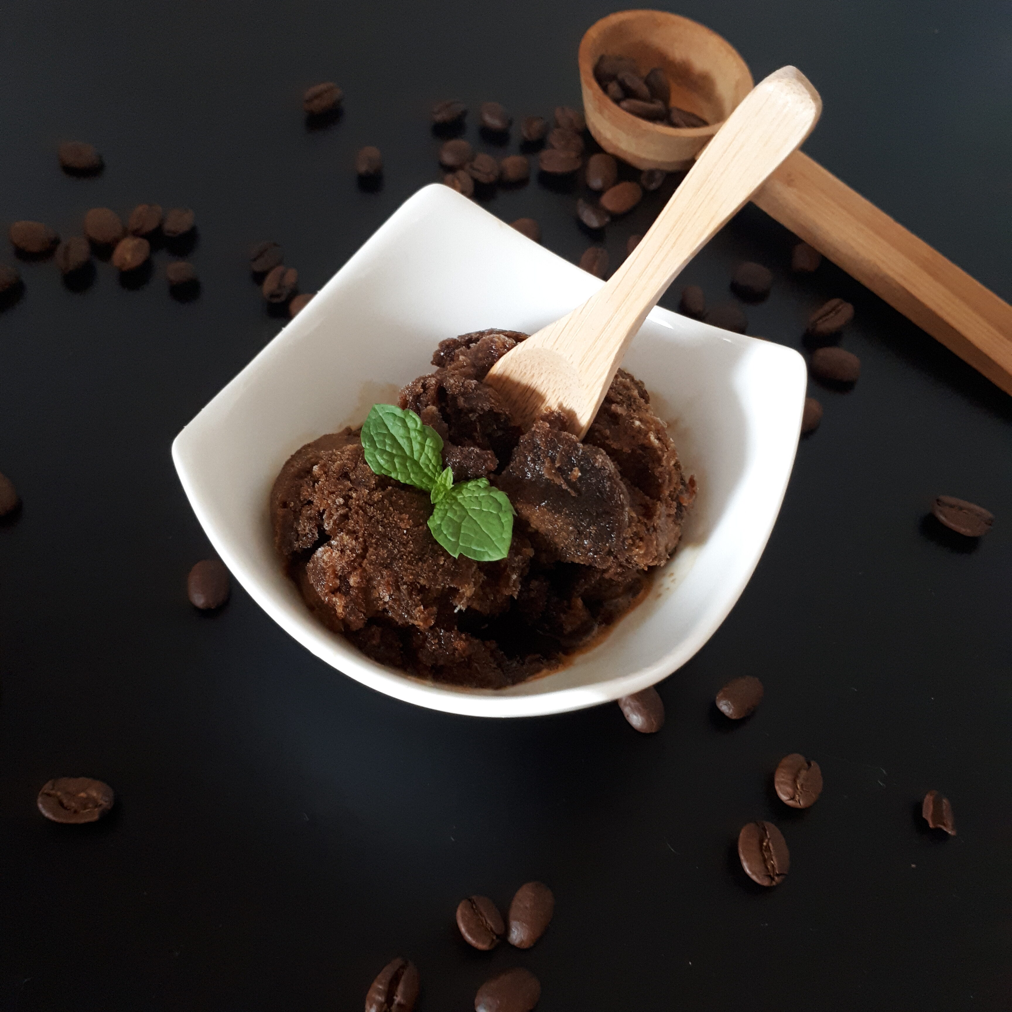 Biela miska s kávovým sorbetom ozdobeným mätou drevená zmrzlinová lyžička okolo rozsypané zrnká kávy v pozadí drevená odmerka na kávu na čiernom povrchu