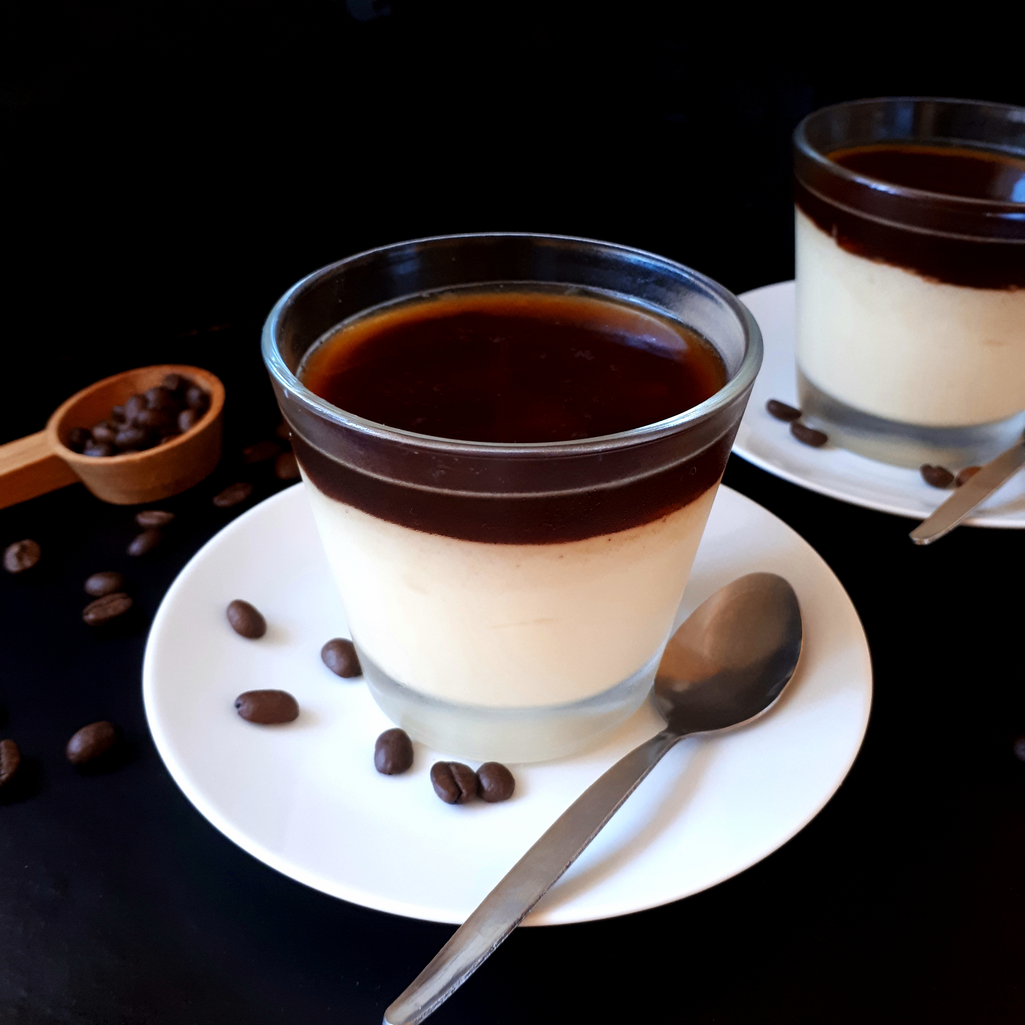Dva poháre s vanikovým pudingom a vrstvou kávovej želatíny na bielych tanierikoch dve lyžičky na čiernom povrchu v pozadí odmerka na kávu okolo rozsypané kávové zrnká