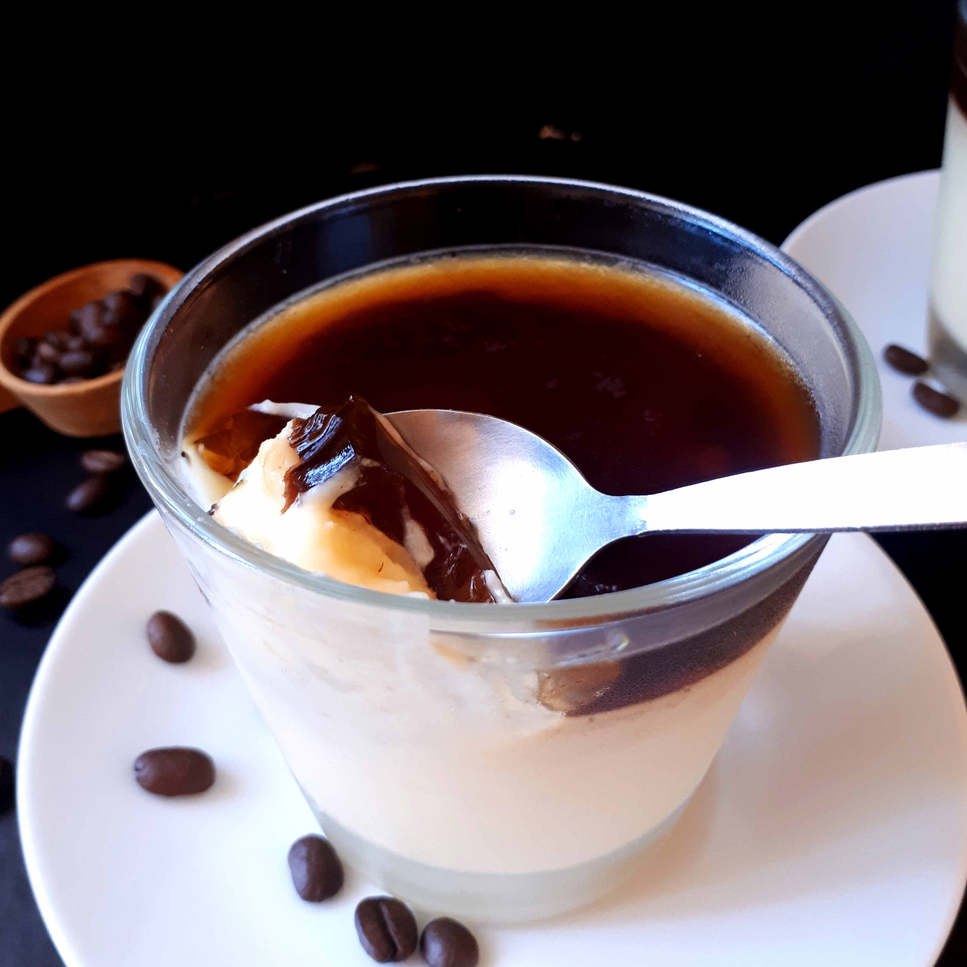 Dva poháre s vanikovým pudingom a vrstvou kávovej želatíny na bielych tanierikoch dve lyžičky na čiernom povrchu v pozadí odmerka na kávu okolo rozsypané kávové zrnká