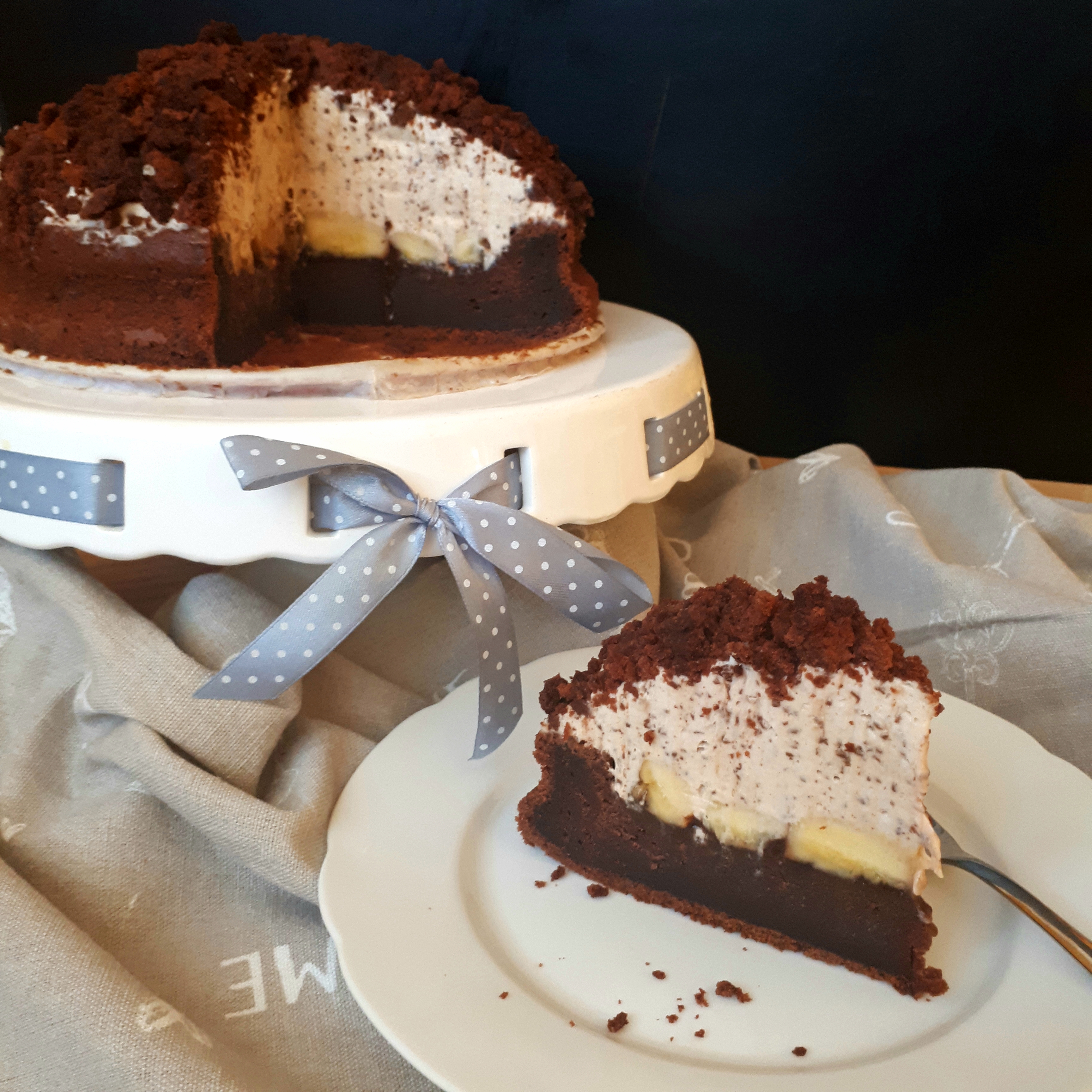 Torta z kakaového cesta a smotanového krému s kúskami čokolády posypanom nadrobeným korpusom na bielom tortovom podnose so šedou mašľou jeden kúsok torty na bielom tanieri dezertná vidlička čierne pozadie a šedý obrus nazbieraný