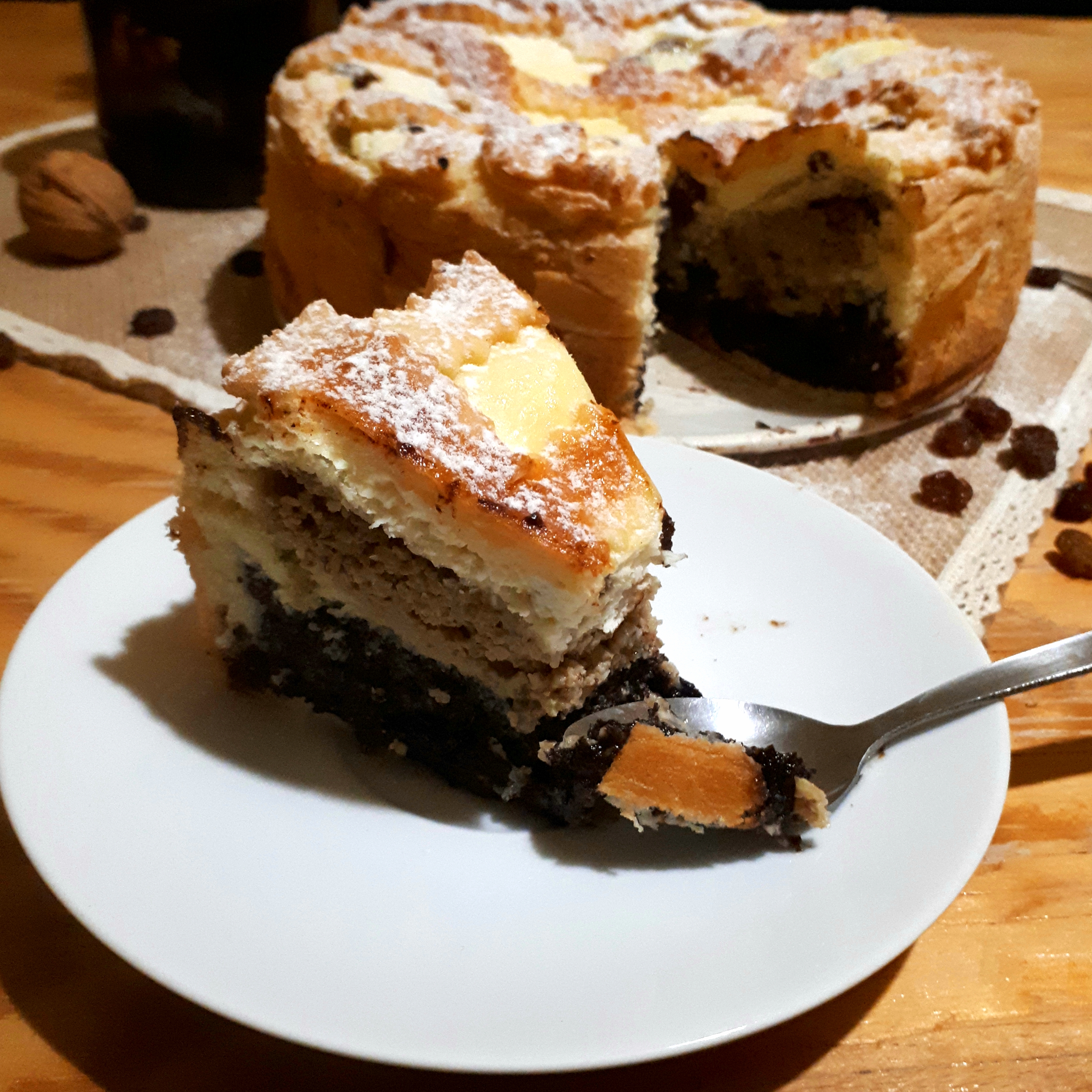 Torta štedrák posypaná práškovým cukrom prírodné filcové prestieranie na drevenom stole okolo hrozienka vlašský orech v pozadí dóza so slivkovým lekvárom v popredí kúsok torty na bielom tanieriku lyžička