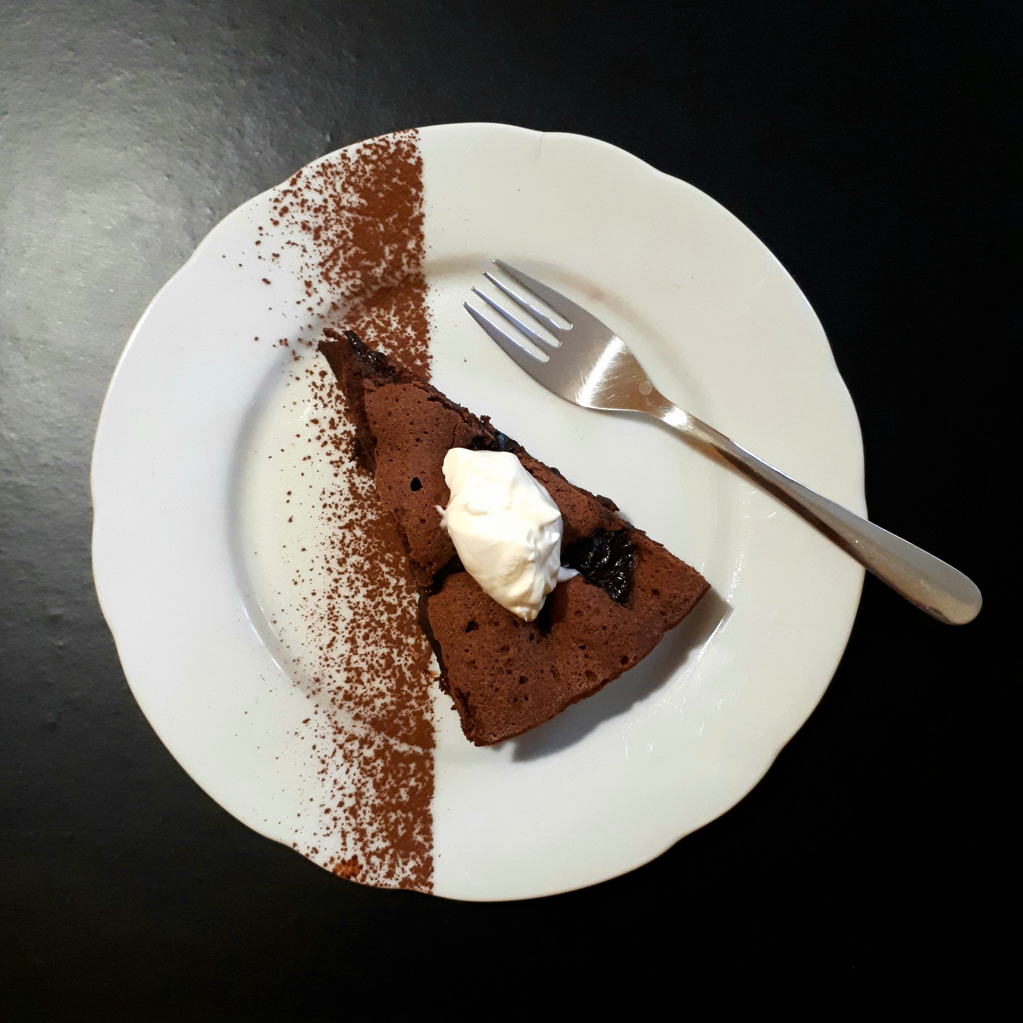 Kúsok čokoládového koláča so sušenými slivkami navrchu kopček kyslej smotany na bielom tanieri posypanom kakaom nerezová dezertná lyžička na čiernom povrchu