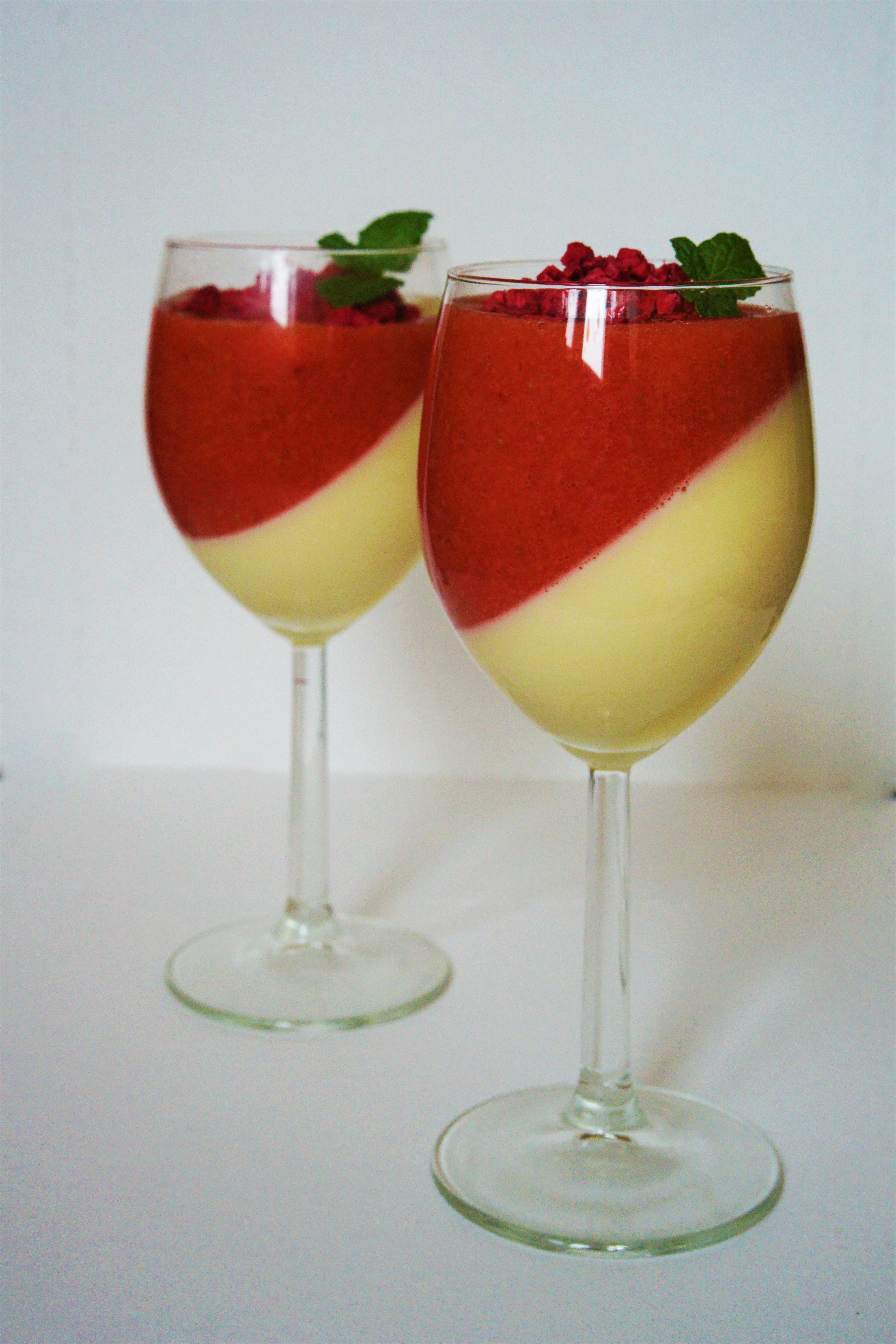 Dva vínové poháre s vrstvou panna cotty a jahodového prelivu dozdobené lyofilizovanými malinami a lístkami mäty biele pozadie