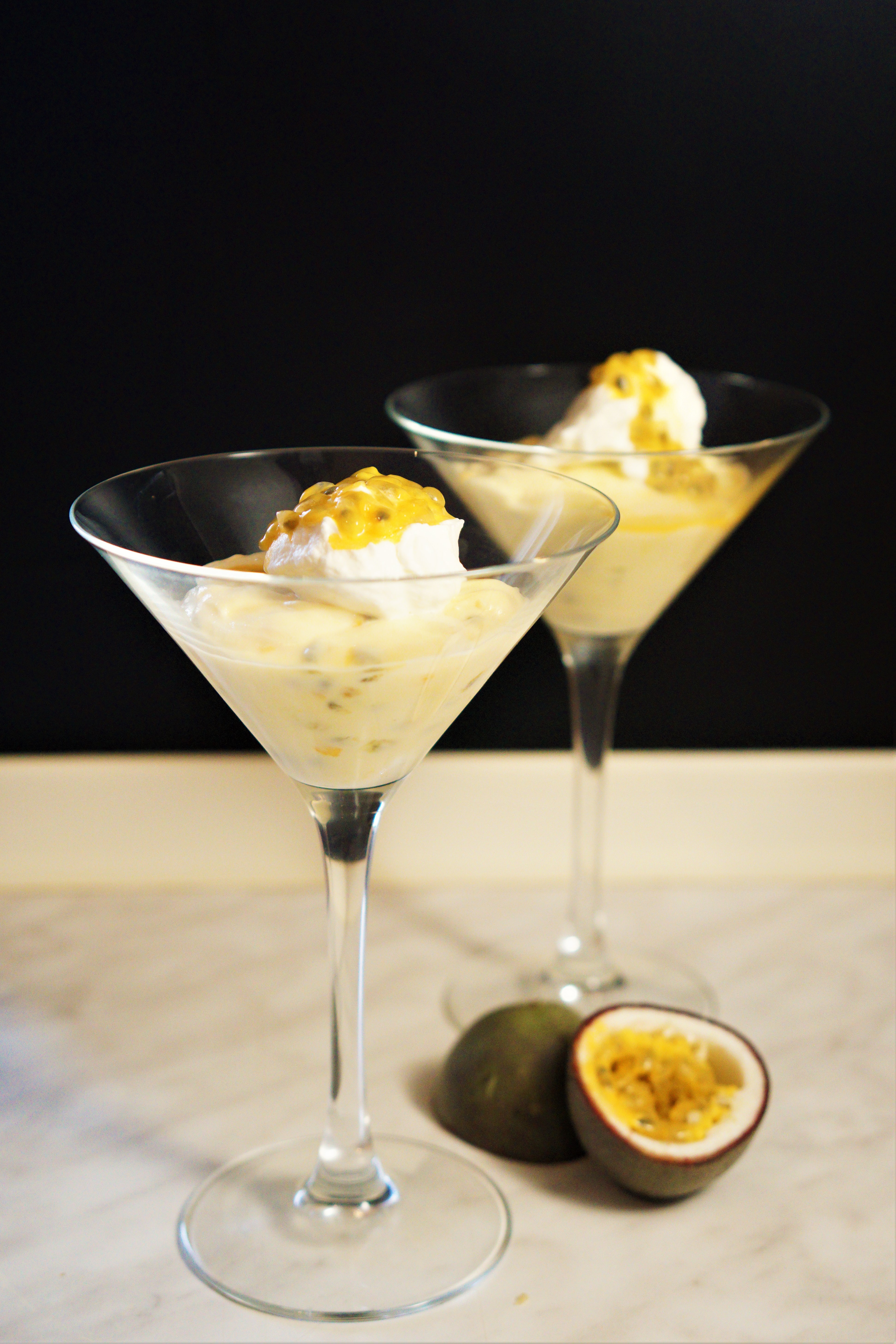Dva poháre na martiny so žlou marakujovou penou kopčekom šľahačky a marakujovou dužinou vedľa prekrojená marakuja na mramorovom povrchu čierne pozadie