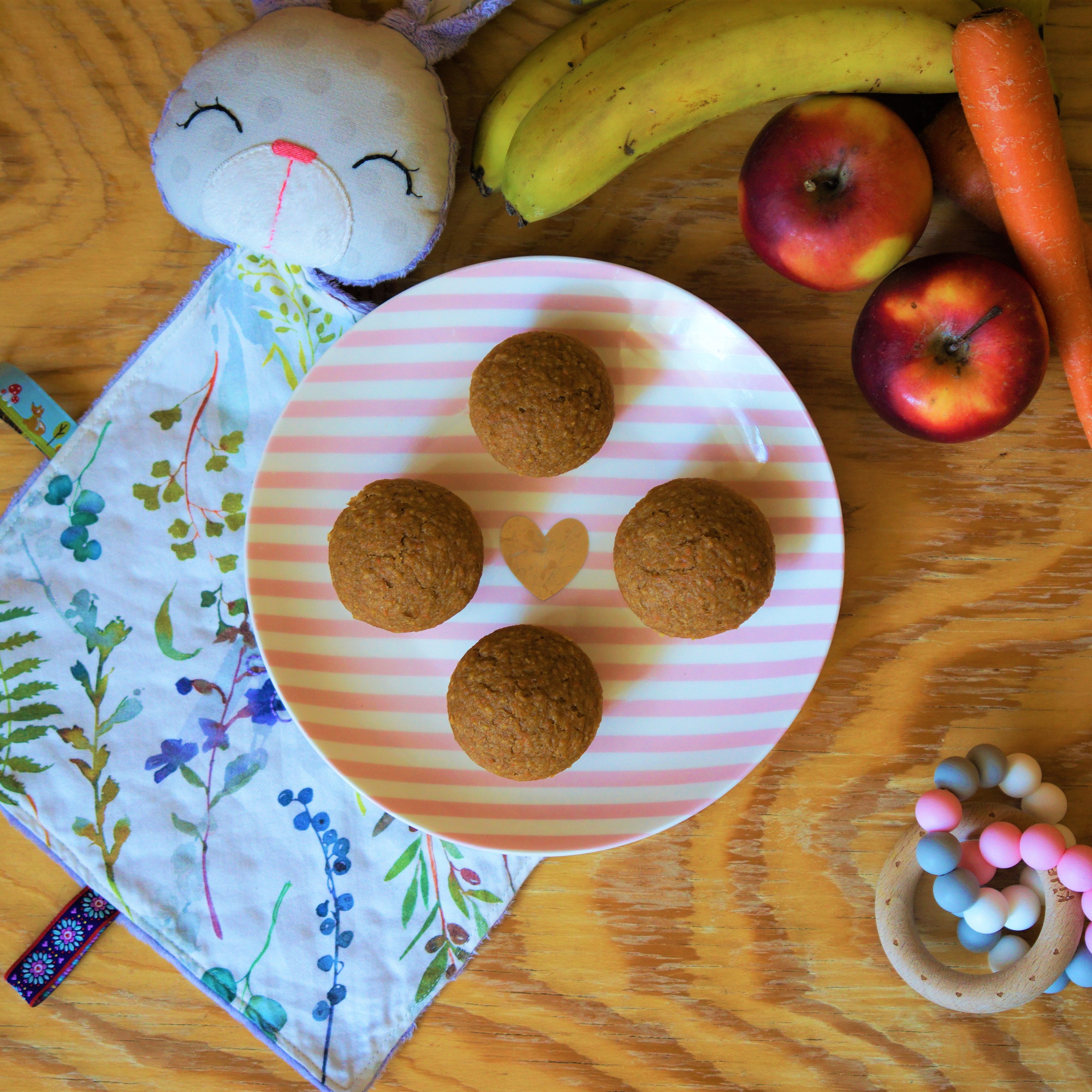 Štyri muffiny na ružovo bielom pásikavom tanieri v strede zlaté srdiečko na drevenom stole fialový látkový zajačik s lúčnymi kvetmi dve jablká dva banány mrkva ružovo šedé kúsatko s dreveným krúžkom
