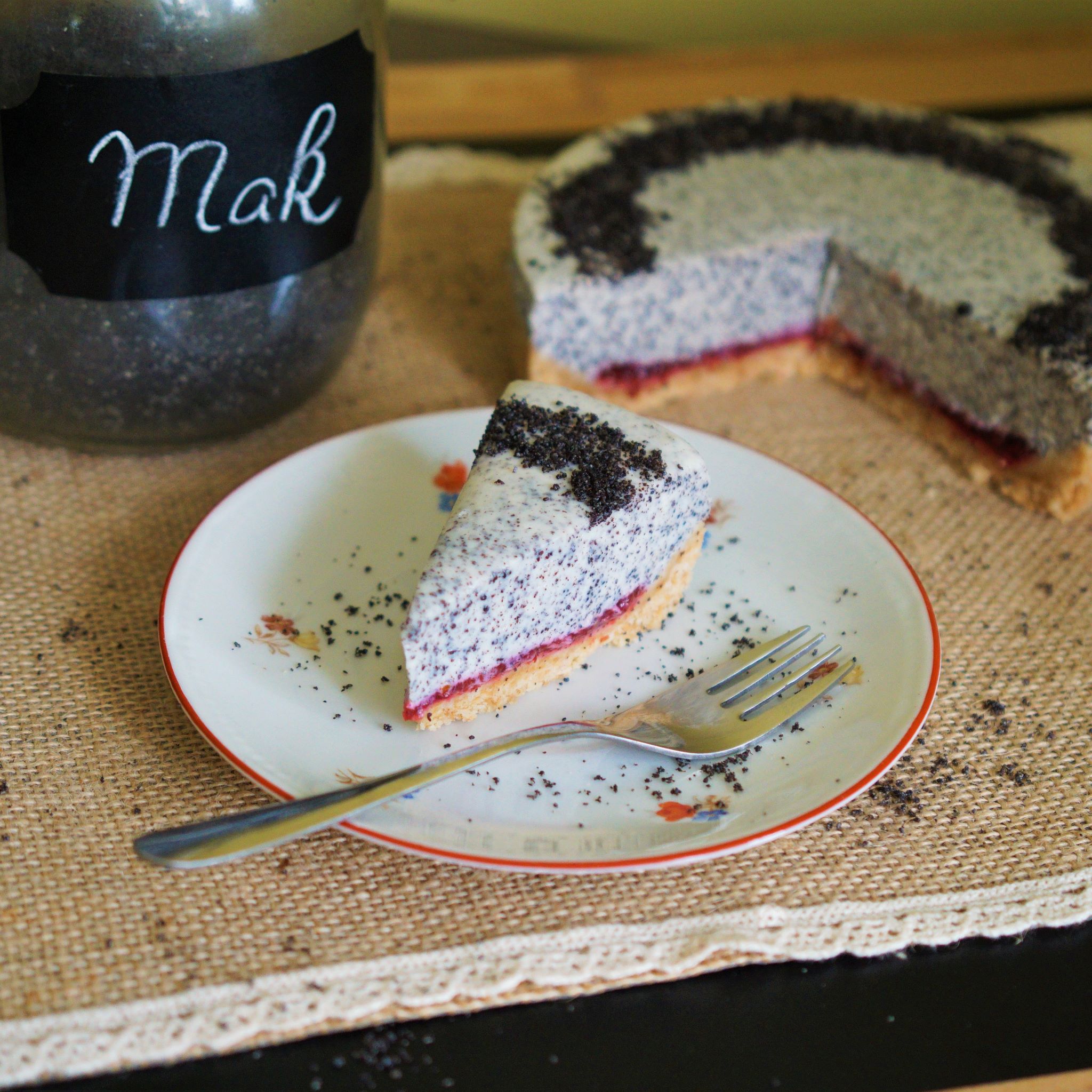 Kúsok makového cheesecake so slivkovou vrstvou na malom bielom tanieriku s červeným okrajom a kvetinkami nerezová dezertná lyžička obsypané mletým makom v pozadí zvyšok cheesecakeu a dúýza s makom na jutovom prestieraní na čiernom povrchu