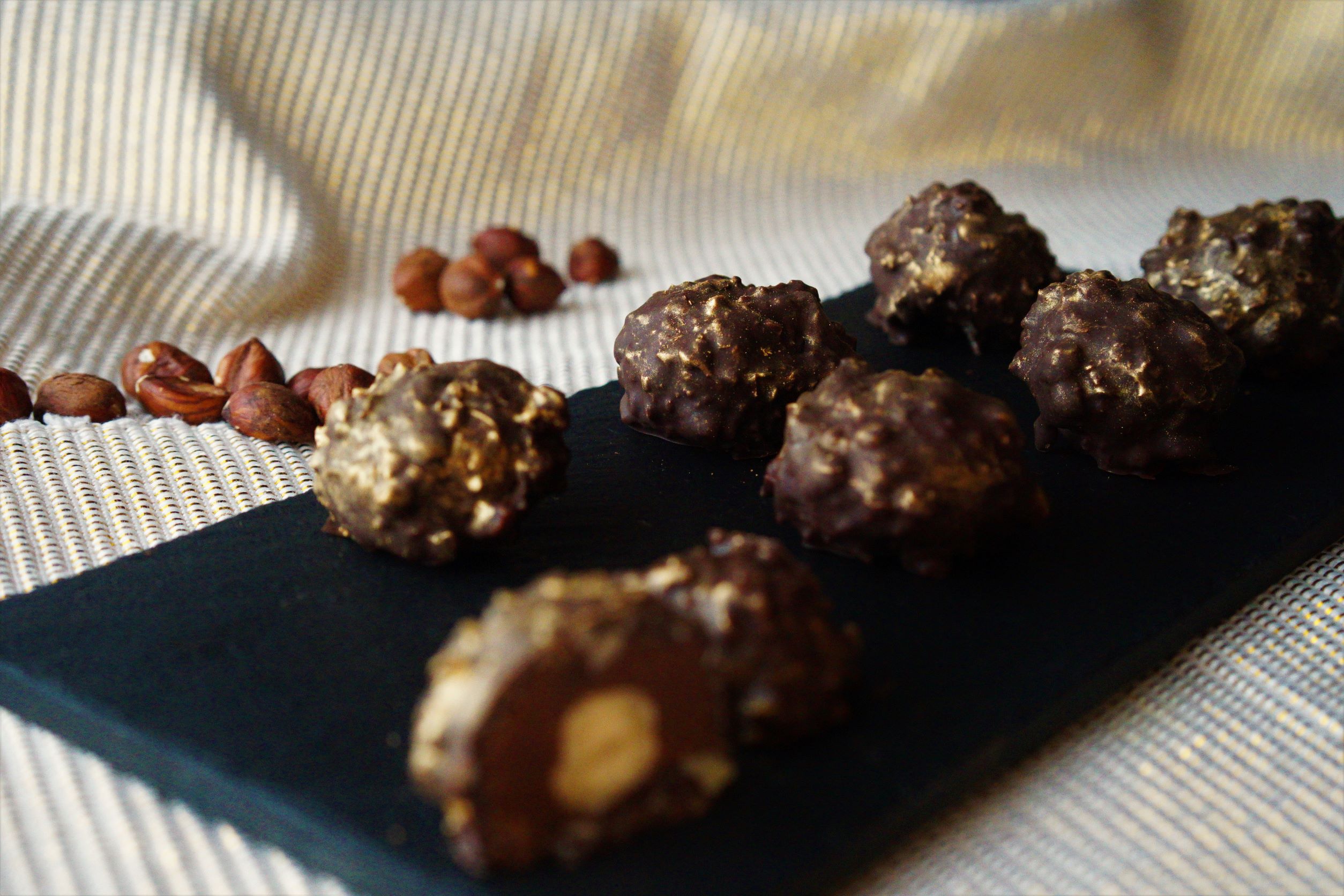 Čierna kamenná doska s čokoládovými pralinkami obalené v lieskových orechoch a zlatom prachu jedna pralinka prerojený vnútro čokoládová praliné a celý lieskový orech ako podklad a pozadie zlatý obrus okolo rozsypané lieskové orechy