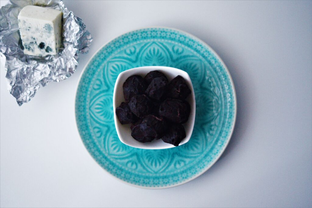 Čokoládové truffles s čiernym kakaom v bielj štvorcovej miske položenej na modrom vzorovanom tanieri biely podklad vedľa syr niva v alobale