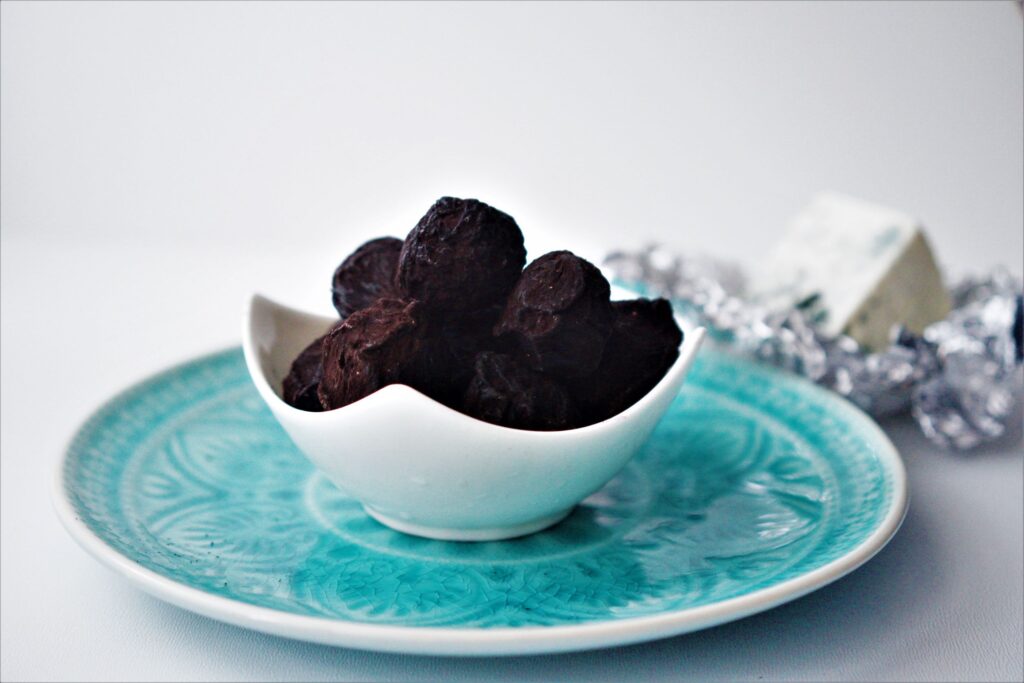 Čokoládové truffles s čiernym kakaom v bielj štvorcovej miske položenej na modrom vzorovanom tanieri biely podklad vedľa syr niva v alobale