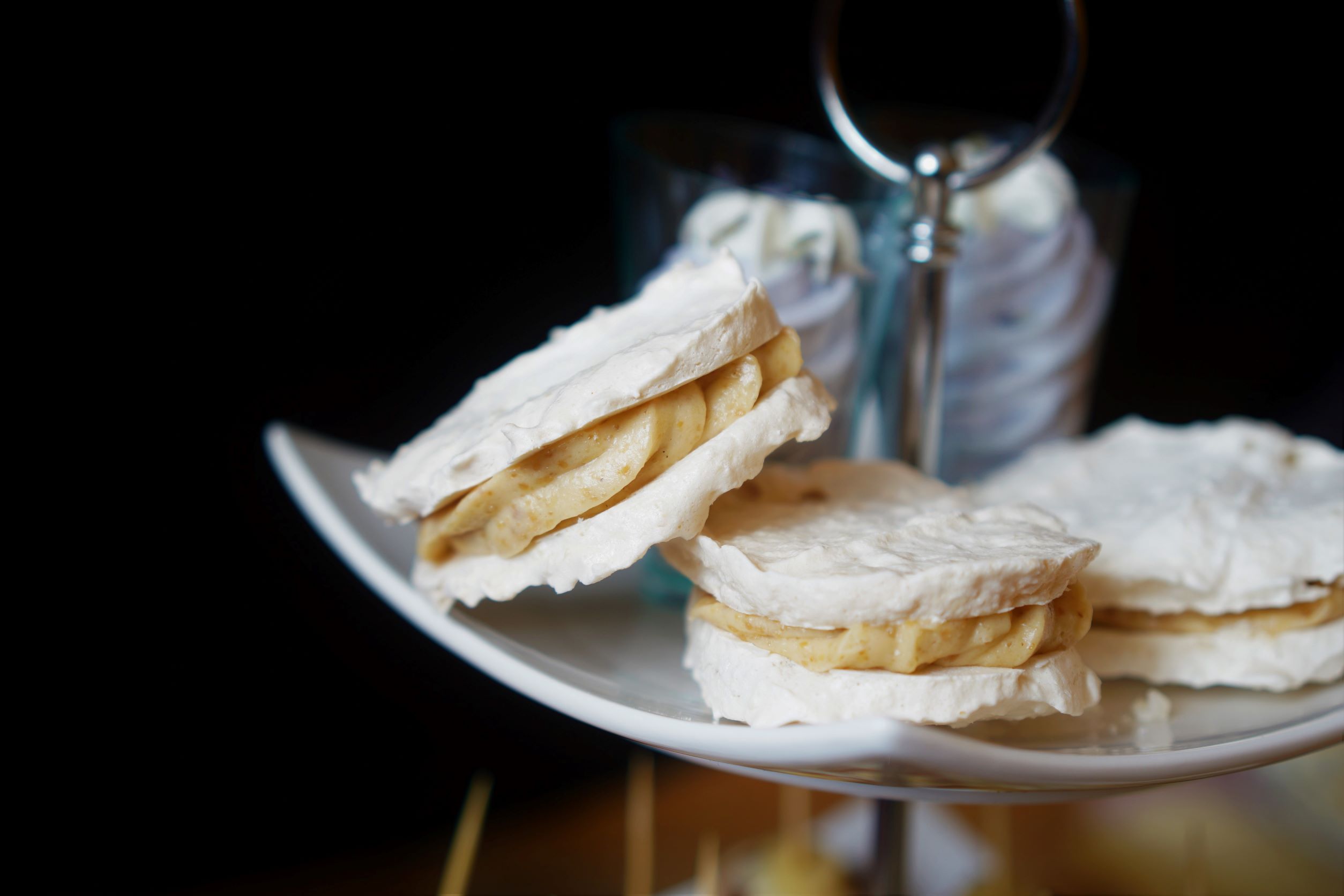 Biely porcelánový etažér a na ňom tri laskonky s grilážovým krémom v pozadí dva poháriky s levanduľovou penou na spodnom tanieri jednohubky čierne pozadie