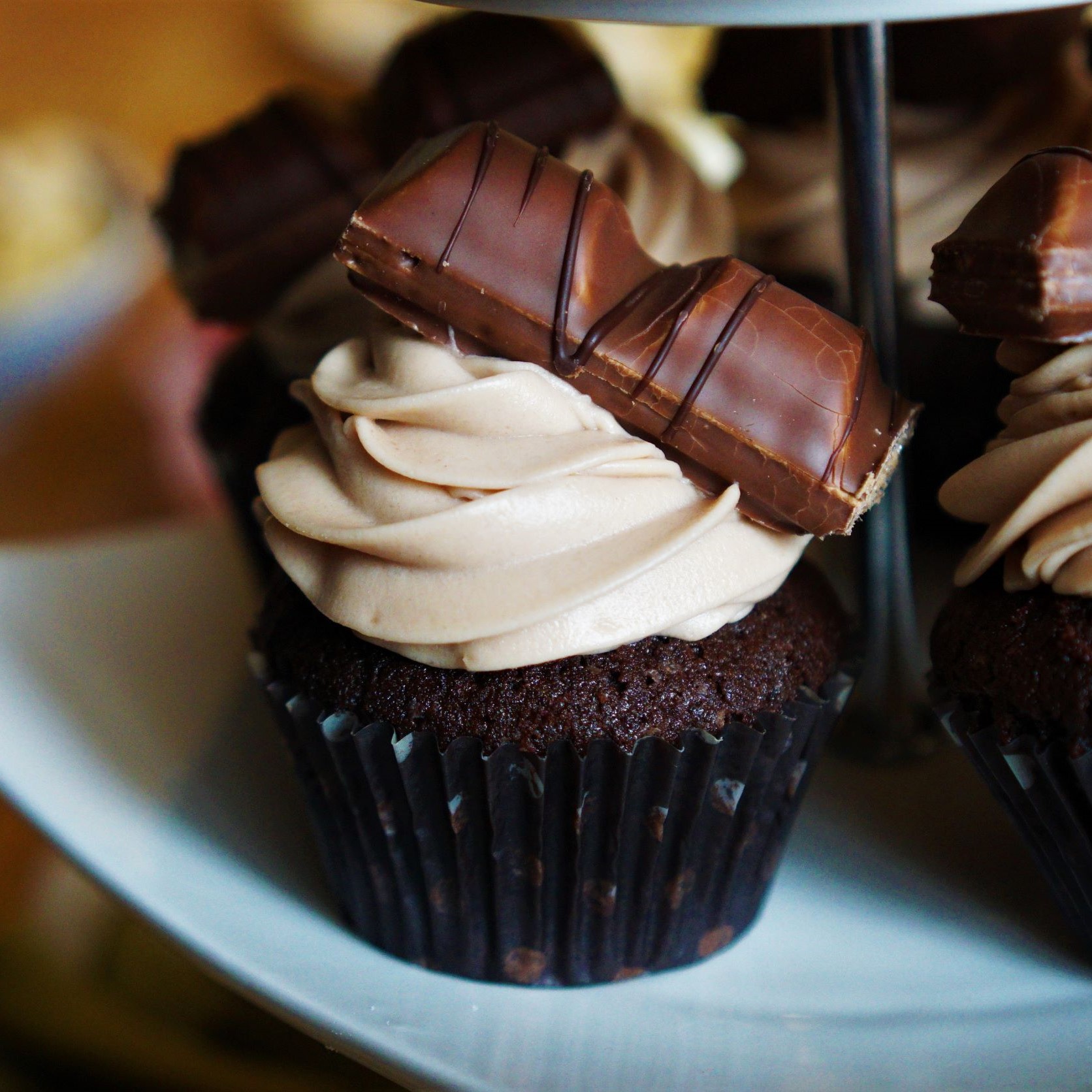 Biely keramický etažér s cupcakes kakaového cesta a krému z kinder čokolády dozdobené tyčinkou kinder bueno v pozadí drevený stôl