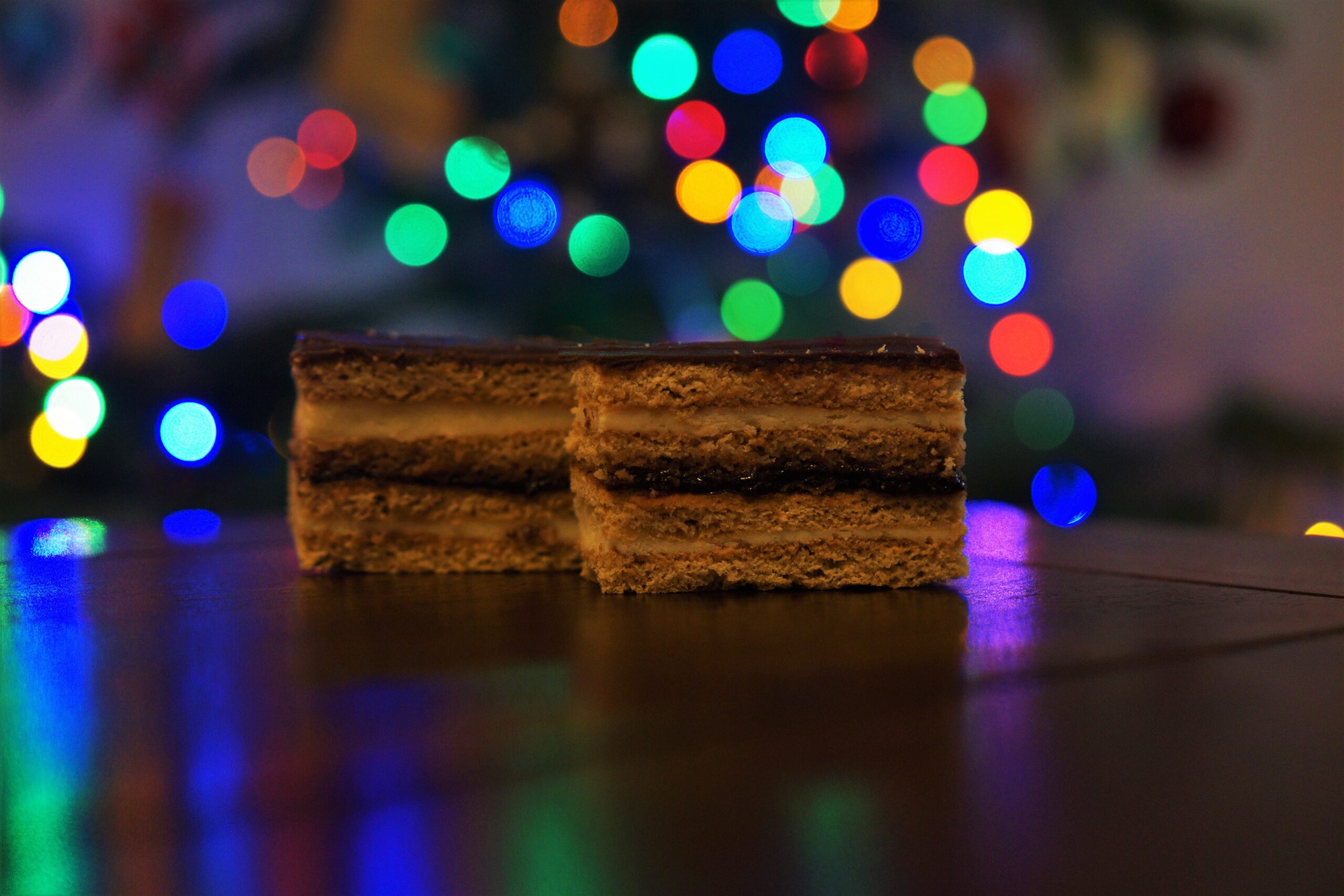 Dva kúsky medového rezu so svetlou náplňou a lekvárom poliate čokoládovou polevou na tmavo drevenom stole v pozadí vianočný stromček