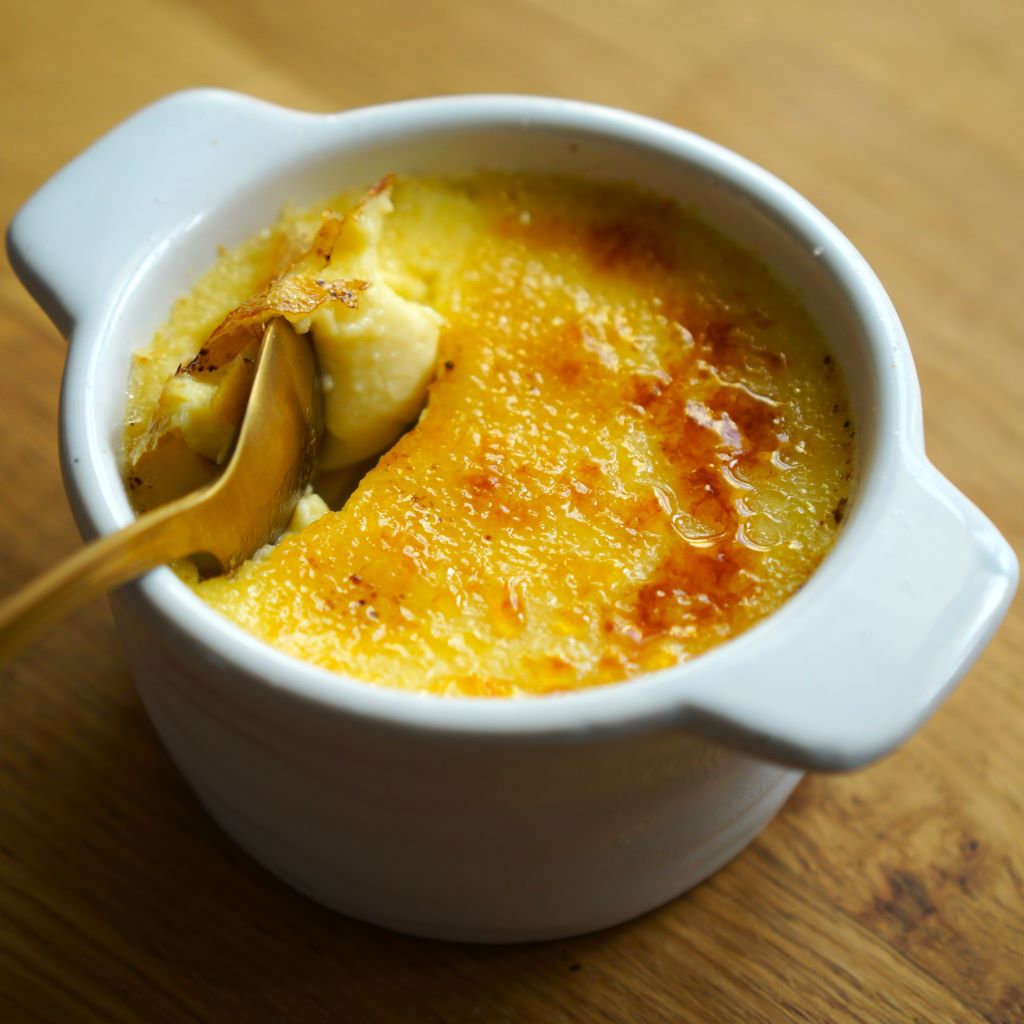 Biela zapekacia miska s creme brulee a zlatou lyžičkou na drevenom stole