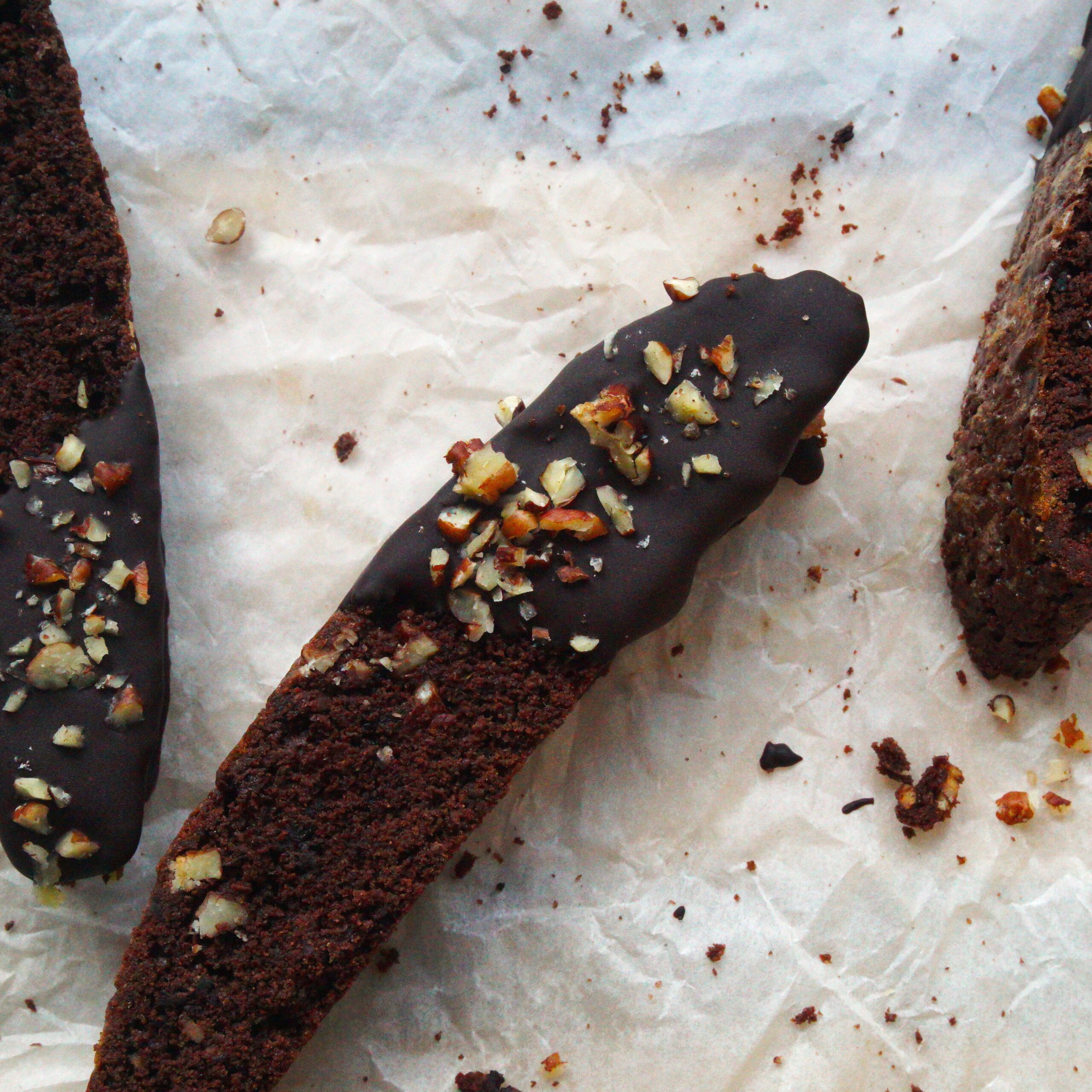 Tri kúsky čokoládových biskotti sušienok polovica namočená v horkej čokoláde a posypaná pekanovými orechami na papieri na pečenie.