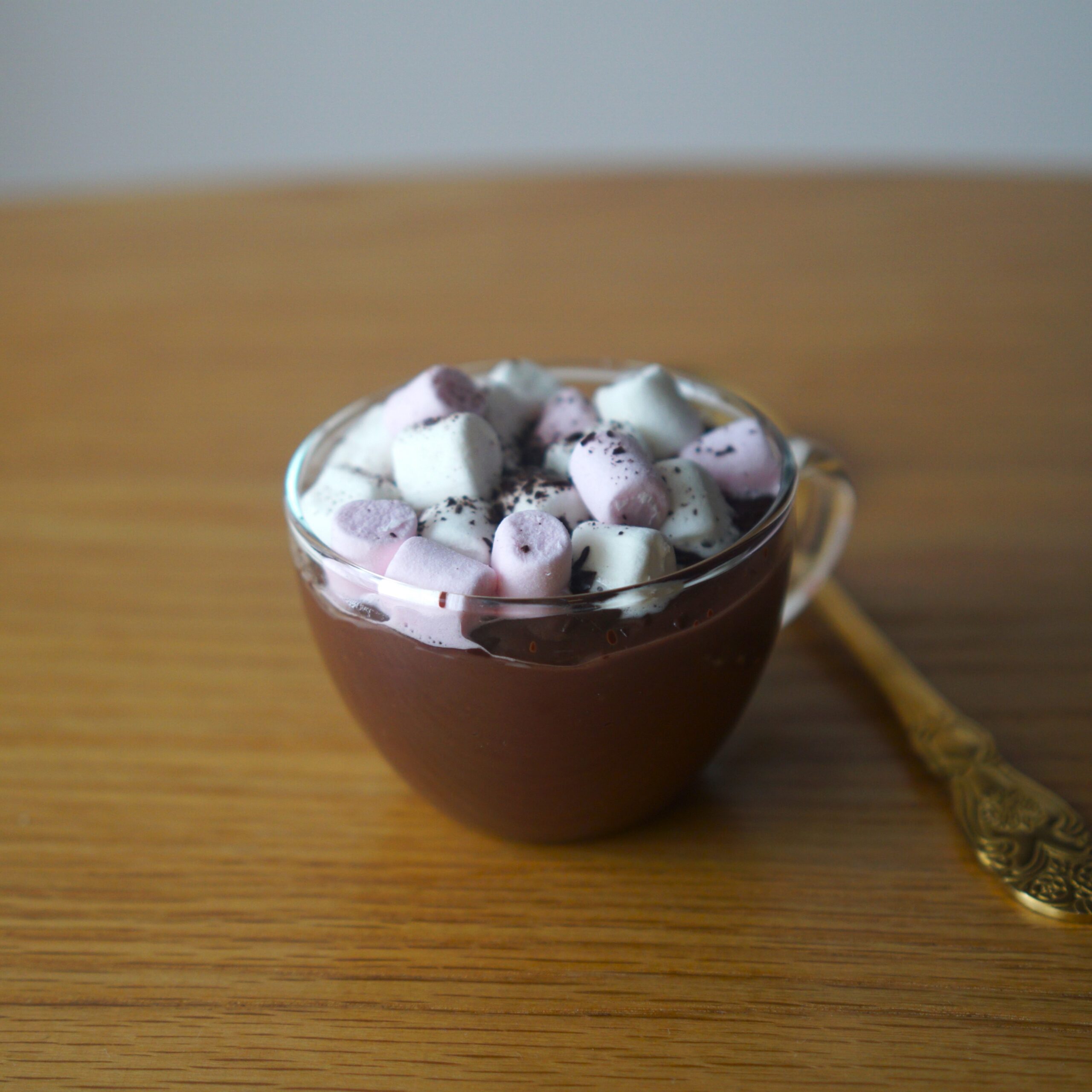 Sklená šálka s horúcou čokoládou posypanou mini marshmallows a nastrúhanou čokoládou zlatá lyžička na drevenom stole biele pozadie